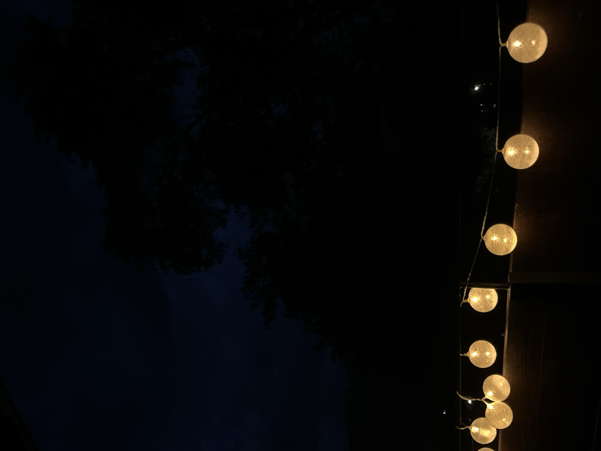 The image depicts a dark outdoor scene at night, likely on a balcony or patio. A string of spherical, illuminated lights hangs horizontally across the lower portion of the frame. These lights are a warm yellow and appear to be made of a textured material, possibly fabric or paper. Behind the lights, a dark, wooden railing or surface is visible. The background is almost entirely black, with the silhouette of tree branches visible against a very dark, night sky. The location is specified as Garmisch-Partenkirchen, Germany. No people are present in the image.
