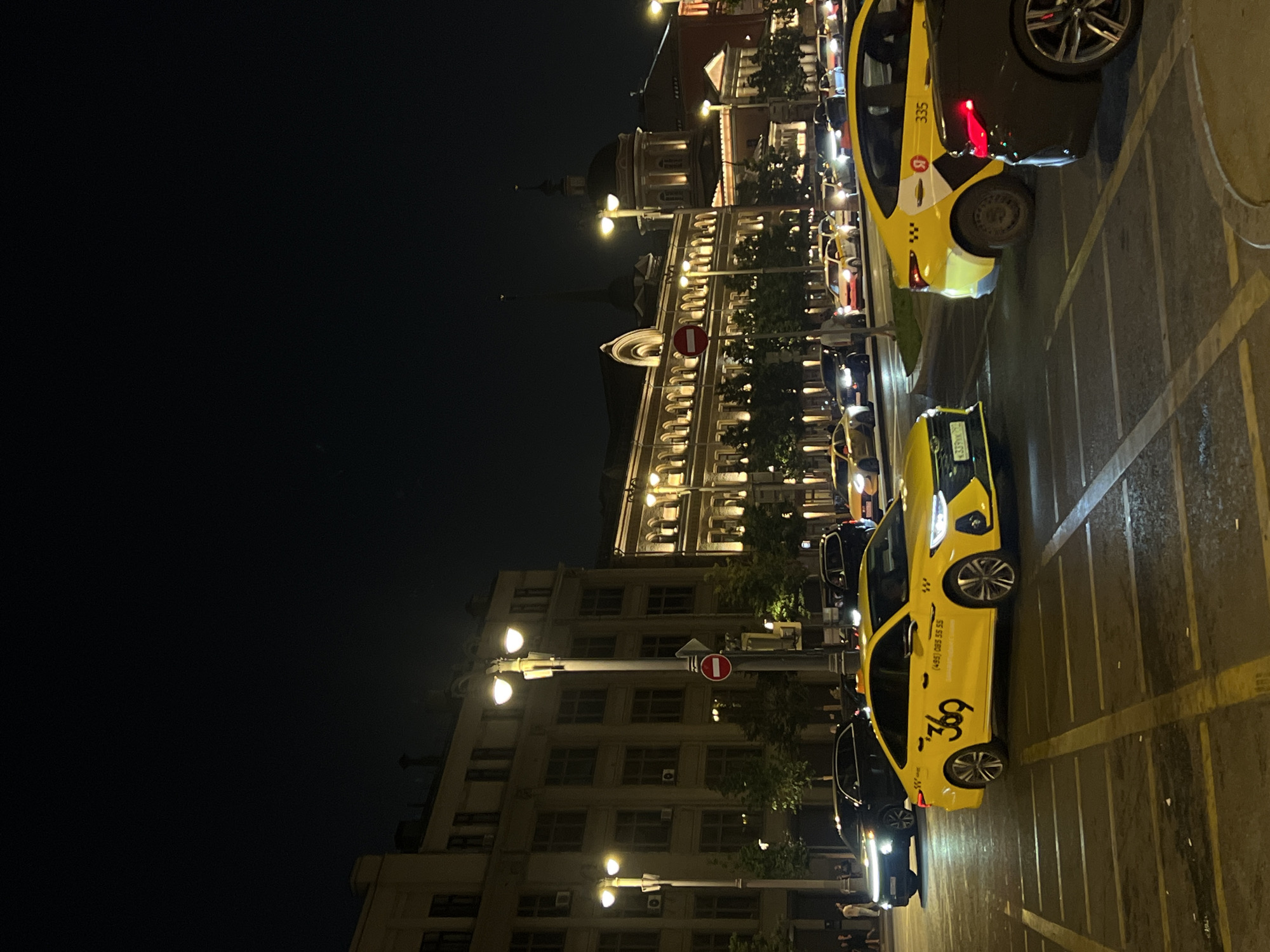 In the city of Unknown, Unknown, at night, a street scene is captured. The sky is dark, with the illumination coming from streetlights and building lights. Several yellow taxis are visible, indicating a busy urban environment. One taxi in the foreground prominently displays the number "369" and a phone number. Other vehicles, including dark-colored cars, are also present on the road. Buildings with multiple windows line the street, some with lights on, suggesting active occupancy. Trees are sparsely planted along the sidewalk. A traffic sign with a red circle and a horizontal bar is mounted on a lamppost. The road surface has painted lane markings. The scene appears to be a street intersection or a busy thoroughfare. There are a few individuals walking on the sidewalk in the background, their faces indistinct due to the distance and low light. The overall impression is that of a typical urban night with traffic and illuminated architecture.