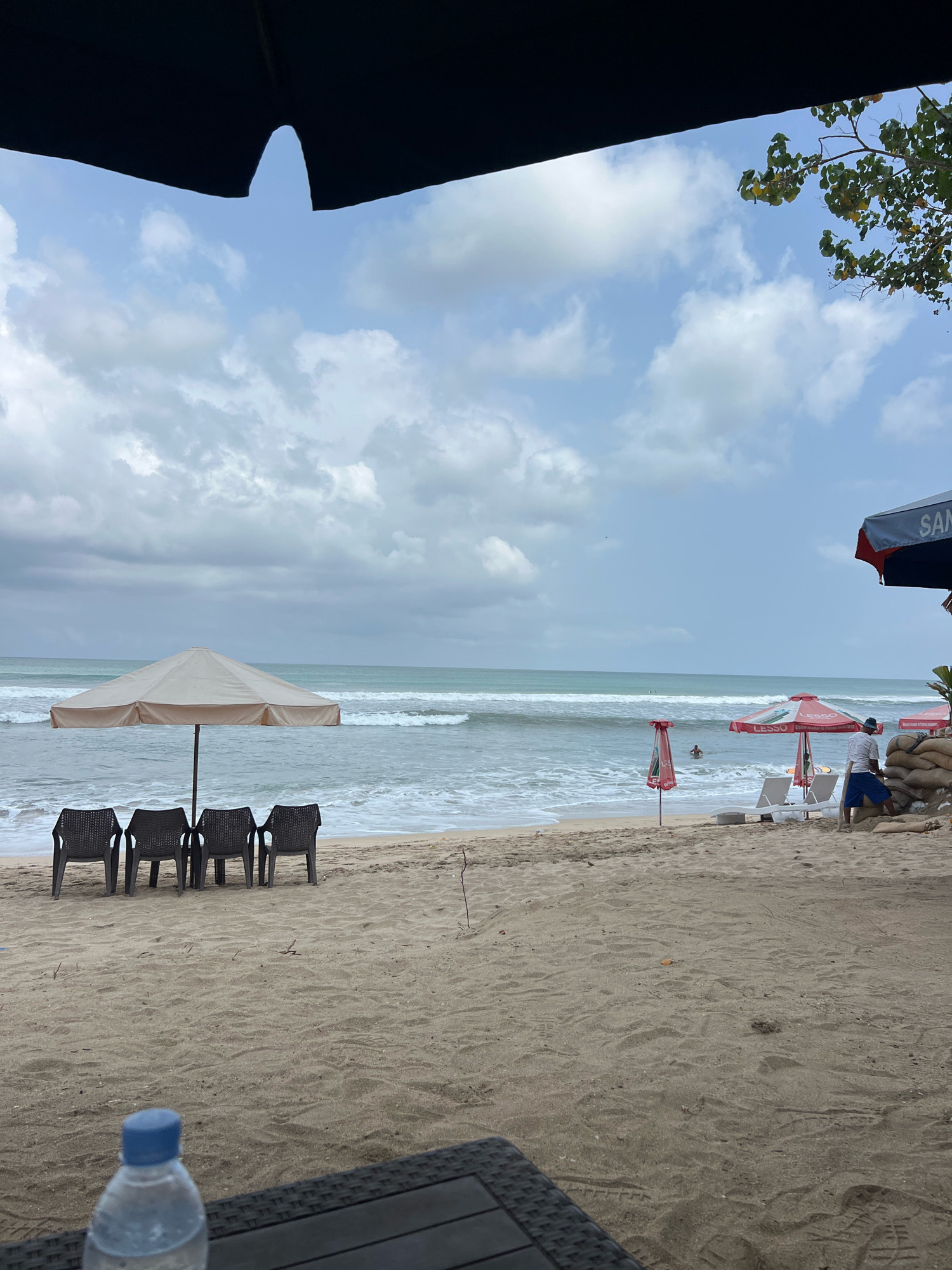 This is a horizontal, eye-level shot of Kuta Beach in Bali, Indonesia, on an overcast day. The foreground is dominated by the sandy beach, with a water bottle and a dark woven surface in the lower left corner. Further into the scene, four empty dark plastic chairs are arranged in a row under a large, light-colored beach umbrella. Beyond them, the ocean stretches out with gentle waves.
On the right side of the frame, a man in blue shorts and a white shirt stands near a pile of sandbags. Two white lounge chairs and a red and white umbrella are also visible in this area. Another umbrella, this one dark blue and red with the word "SANT" partially visible, is positioned further to the right, partially obscuring the view of the beach. In the middle distance, a person is swimming in the ocean. The sky is filled with large, fluffy clouds, and a tree branch with green and yellow leaves hangs from the top right corner of the image. The overall impression is of a quiet, possibly post-rain, moment on a tropical beach.