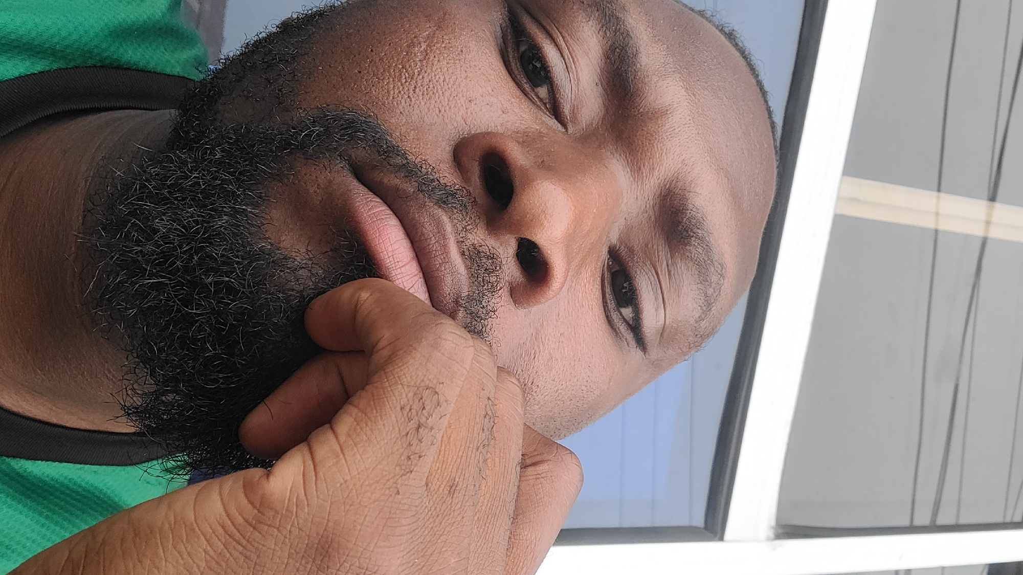 A close-up portrait of a man of African descent is captured with a diagonal tilt. He has dark skin and a full, neatly trimmed black beard. His gaze is directed towards the viewer, appearing calm and somewhat contemplative. He is wearing a green shirt with a black collar. His right hand is raised, with fingers resting near his chin and partially obscured by his beard, reinforcing a pensive or thoughtful pose. The background features a window or glass partition with light-colored frames, through which a soft blue sky and some faint, dark vertical lines (possibly wires or reflections) are visible. The natural, bright lighting suggests it is daytime. No text is visible in the image. The setting appears to be a casual indoor or semi-outdoor space in Jos, Nigeria.