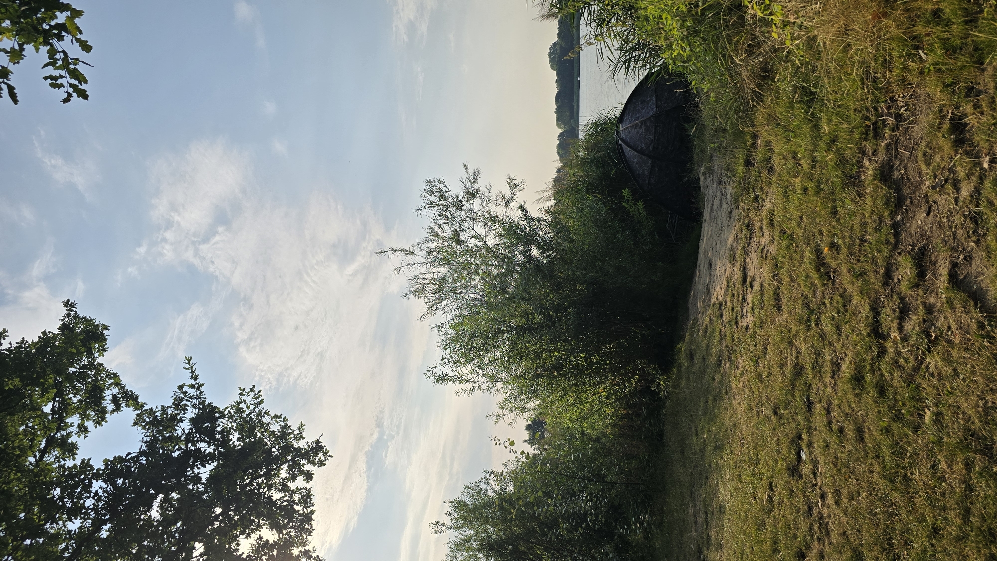 The image depicts a serene lakeside scene, likely at a fishing spot near Kilder, Netherlands. In the foreground, a grassy area with patches of dry earth leads towards a dense thicket of green bushes and tall reeds. Partially obscured by the vegetation, a dark, dome-shaped fishing tent is set up on the bank. Beyond the foliage, a calm body of water stretches out, bordered by a treeline in the distance. The sky above is a pale blue, dotted with wispy white clouds, and the warm light suggests it is either early morning or late afternoon. The overall impression is one of tranquility and outdoor recreation.
