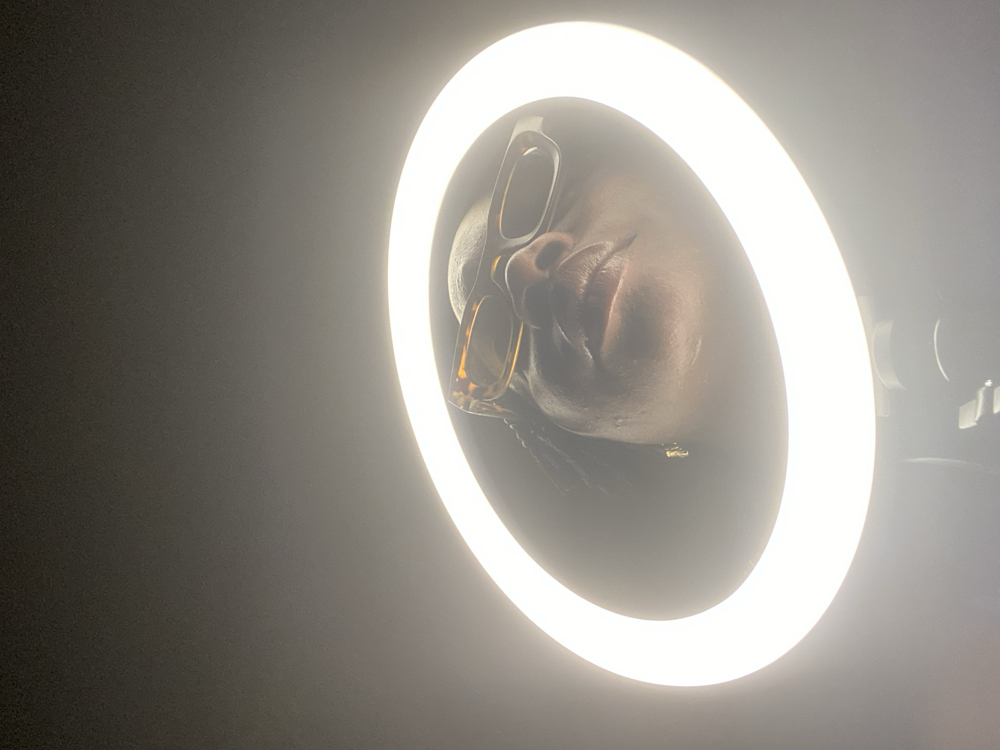 A person's face is centered within a bright circular ring light. The person is wearing glasses with a brown and tan frame. The background is dark. The location is not discernible from the image.