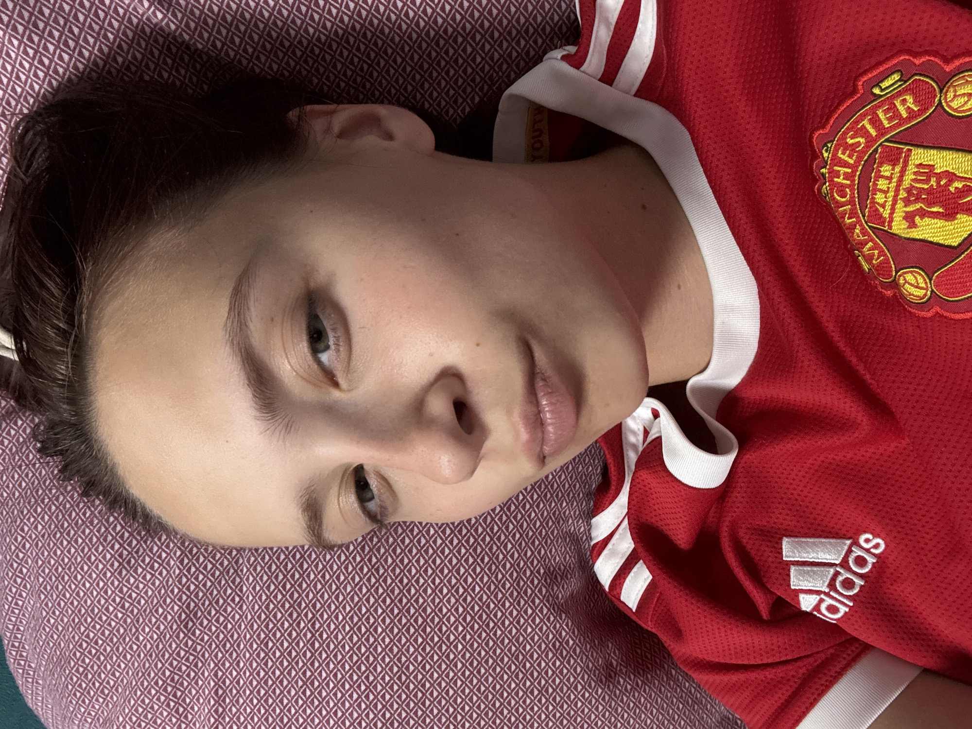 A young woman is lying down, looking at the camera. She is wearing a red Manchester United jersey with white trim and the Adidas logo. Her hair is dark brown and pulled back. The background is a textured, burgundy-colored surface, possibly a pillow or blanket. The lighting appears to be indoor. The image is a close-up shot, focusing on the woman's face and upper body. There is no visible indication of the location being in the city of Прикамский, Russia.