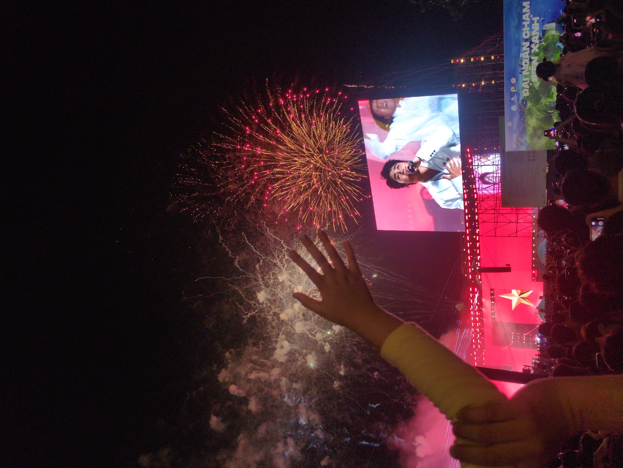 The image depicts a nighttime outdoor event in South Quy Nhon Ward, Vietnam. In the immediate foreground, a human left arm and open hand, with a light-colored sleeve, are raised upwards from the lower left. Below the hand, a dense crowd of attendees occupies the lower third of the frame, with numerous individuals holding up illuminated smartphones. Above the crowd, a large concert stage is visible, illuminated primarily with red lighting, featuring a prominent golden five-pointed star emblem on its backdrop. Centered above the stage is a large rectangular LED screen displaying a male singer with dark hair and a light-colored shirt, holding a microphone and gesturing with his hand over his chest. A second partially visible individual in a white shirt and a straw hat is behind him on the screen. Below the LED screen, a banner with green landscape imagery and Vietnamese text, including "ĐẠI NGÀN CHÀM ĐIỆN XANH," is displayed. The night sky in the background is punctuated by active fireworks, with a large spherical burst of red and yellow sparks visible in the upper right, and white and red smoke trails from other explosions to the left.