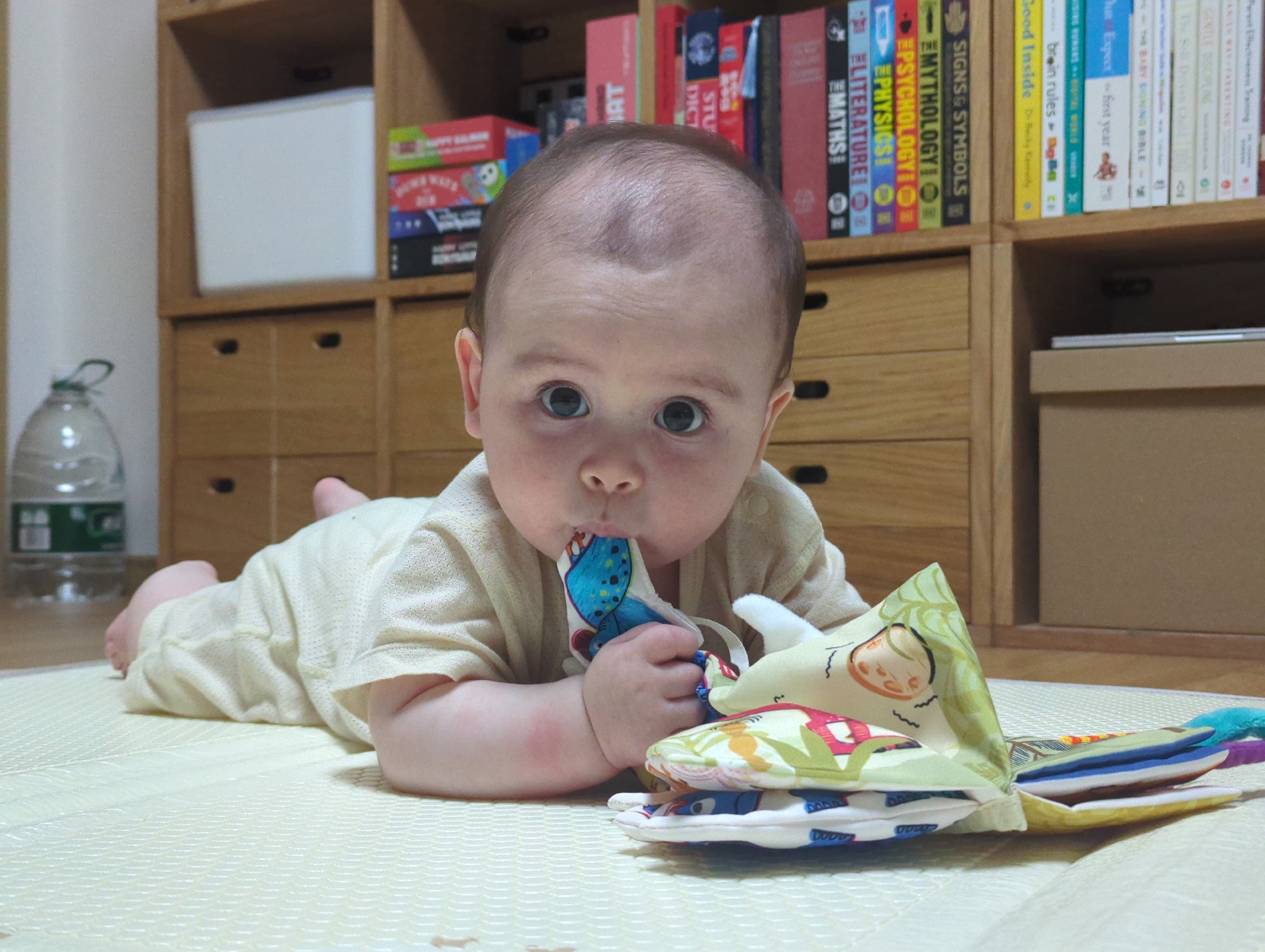 A baby lies prone on a quilted mat, sucking on the corner of a fabric book. The baby is fair-skinned with dark hair and is wearing a light-colored shirt. The book is open, revealing pages of various colors and designs. Behind the baby, a wooden cabinet is visible. The cabinet contains books, including titles like "MYTHOLOGY," "SIGNS & SYMBOLS," "LITERATURE," and "MATHS." A large water jug sits on the floor to the left of the cabinet. The photo appears to have been taken indoors. There is no way to confirm the location is Shunde District, China from the given information.