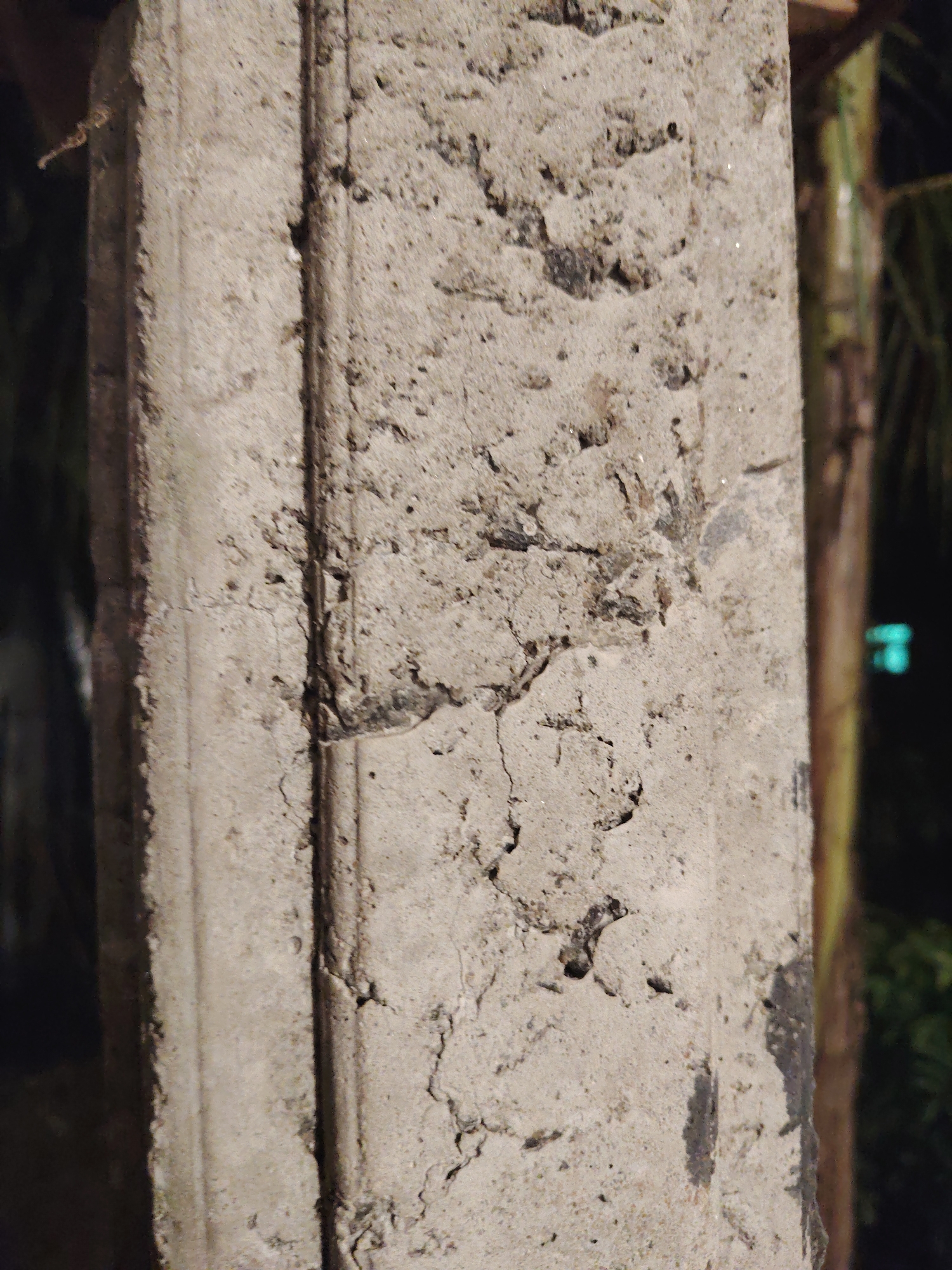This media file features a close-up shot of a textured concrete pillar, positioned vertically and dominating the frame. The pillar is made of light grey concrete, showing significant signs of wear and age, including numerous cracks, rough patches, and darker discolored areas that suggest dirt, mold, or chipped material. A distinct, narrow, linear indentation runs vertically along the left side of the pillar, with a similar, slightly wider indentation further to the right, adding a subtle architectural detail.
The lighting is dim, suggesting it might be evening or night, or the object is situated in a poorly lit environment. The background is heavily out of focus, appearing as blurred dark shapes with hints of green, possibly indicating foliage or distant trees. In some cropped views, a faint, blurred green or blue light source is visible in the far background, reinforcing the idea of a nocturnal or low-light setting. No specific activity or event is taking place, as the image focuses solely on the static structure of the pillar. There is no visible text or identifiable human presence. The overall impression is one of a weathered, utilitarian structure in an indistinct outdoor location.