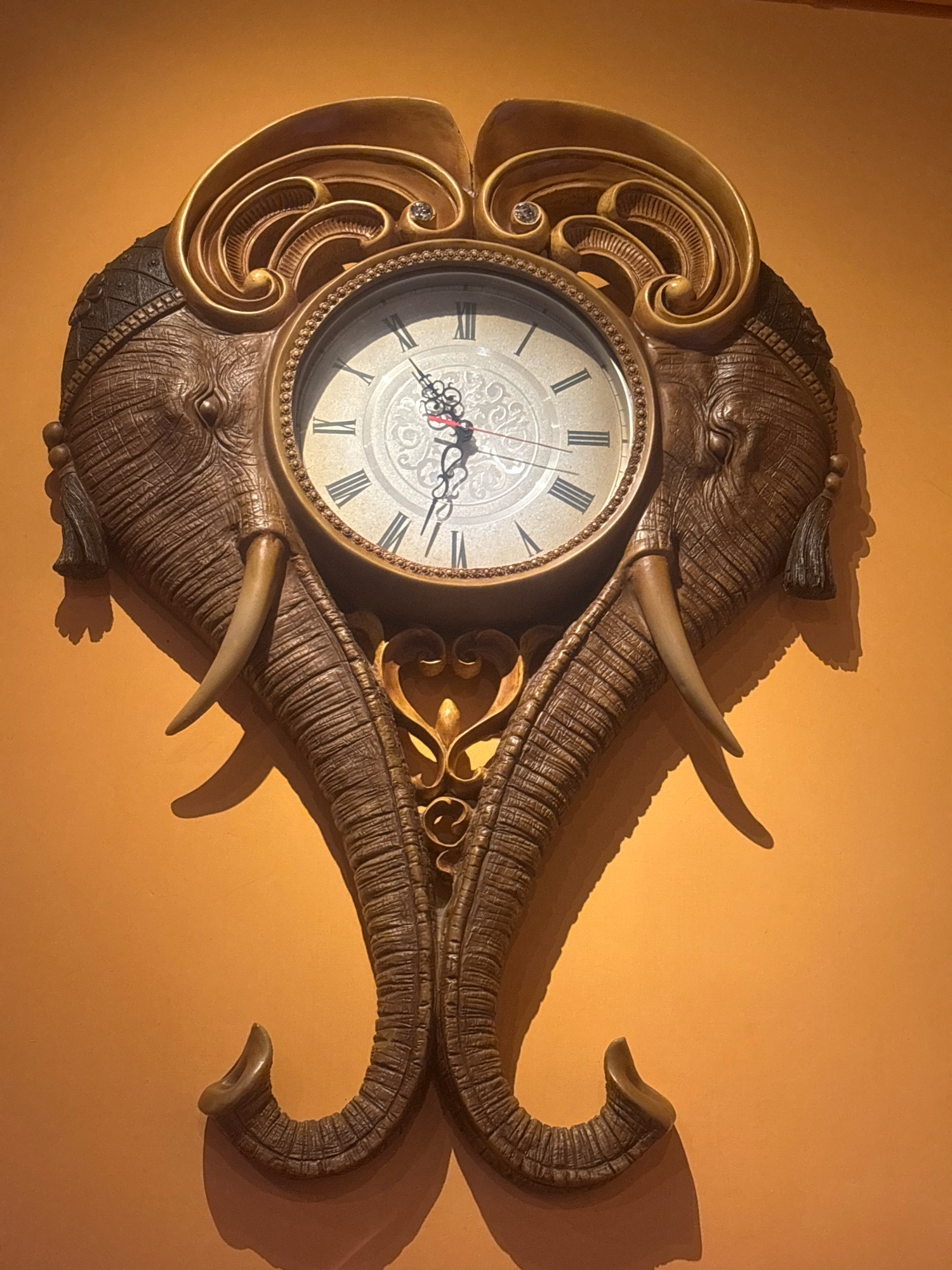 An ornate wall clock, designed in the shape of two elephant heads facing outward, is mounted on an orange wall. The clock face, with Roman numerals and intricate detailing, is positioned centrally between the elephant heads. The elephants are depicted with textured skin, tusks, and decorative tassels hanging from their ears. The overall design is elaborate and appears to be made of a heavy material, possibly resin or a metallic alloy, with a golden finish on the decorative elements. The lighting casts shadows on the wall, indicating a single light source from above and to the left. There are no people or other objects visible in the frame. The clock appears to be set to approximately 3:07. Without further context, its specific location within Macau, China, cannot be determined.