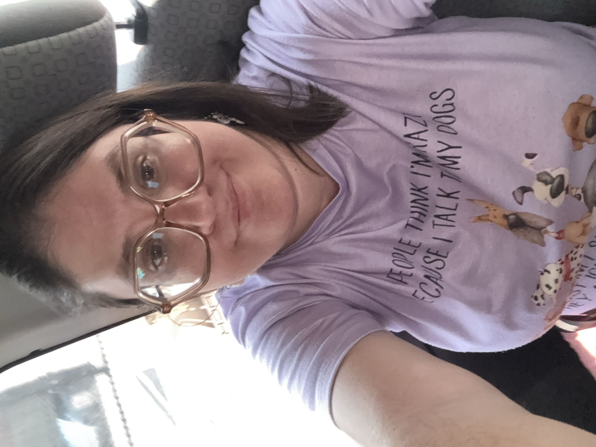 A selfie depicts a woman with dark hair and glasses, wearing a light purple t-shirt with dog images and the phrase "People think I'm amazing because I talk to my dogs." The background includes the interior of a vehicle, with a gray patterned seat visible. No information in the image definitively places the subject in Jiutepec, Mexico.
