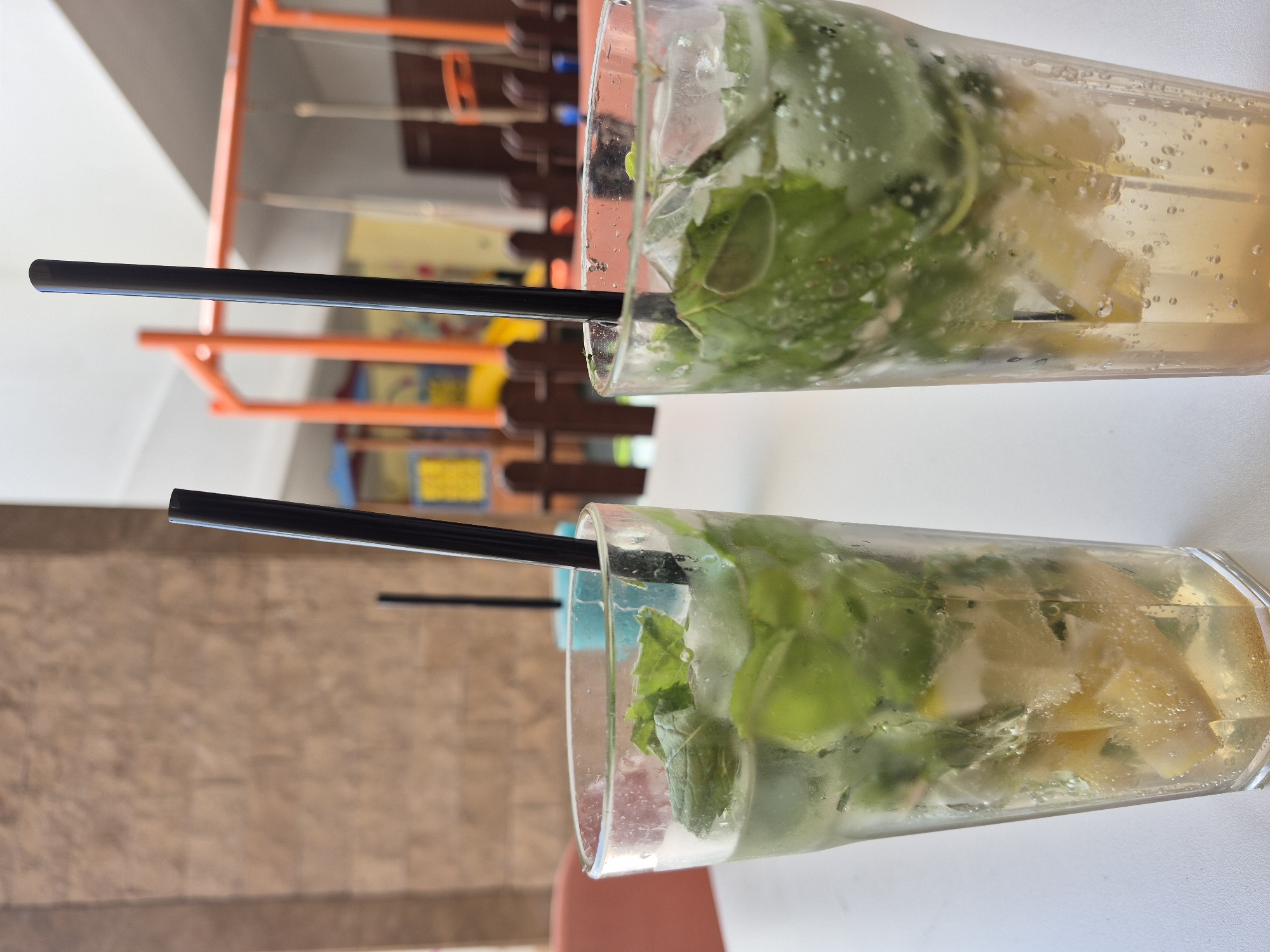 The image shows two mojito cocktails on a white table. Each drink contains mint leaves, lemon or lime slices, and bubbles, with a black straw in each glass. The background is blurred, but visible elements suggest the location might be in Alanya, Turkey, based on the style of outdoor furniture and color palette. There are no people explicitly visible in the image, although there is outdoor patio style furniture.