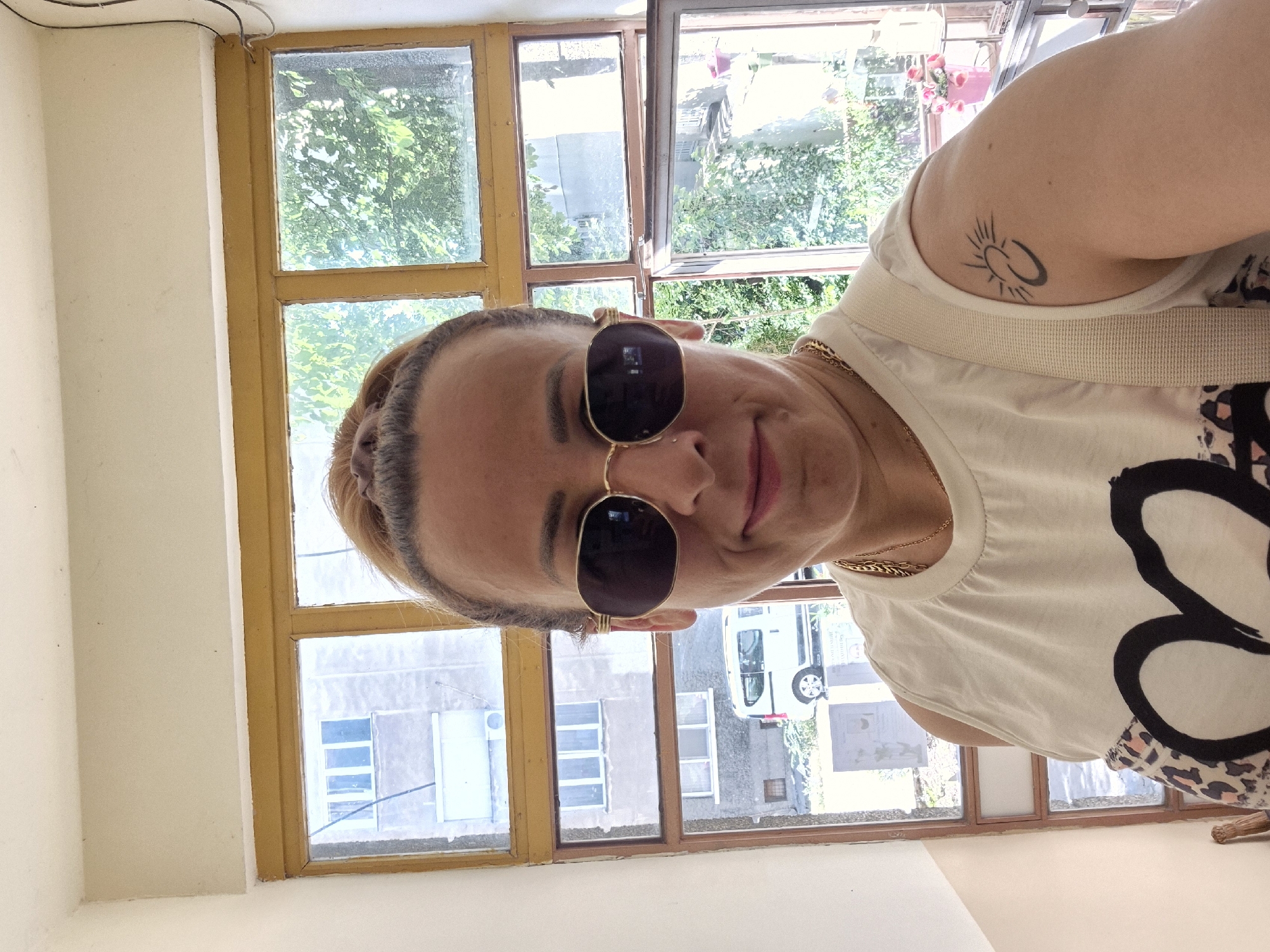 A woman with blonde hair pulled into a bun, wearing sunglasses, a white tank top with a large black graphic, and a leopard print skirt, takes a selfie in a stairwell. She is positioned in front of a multi-paned window that looks out onto a city scene with buildings and trees. A white van is visible parked outside. She has a tattoo of a sun with a crescent moon on her left arm.