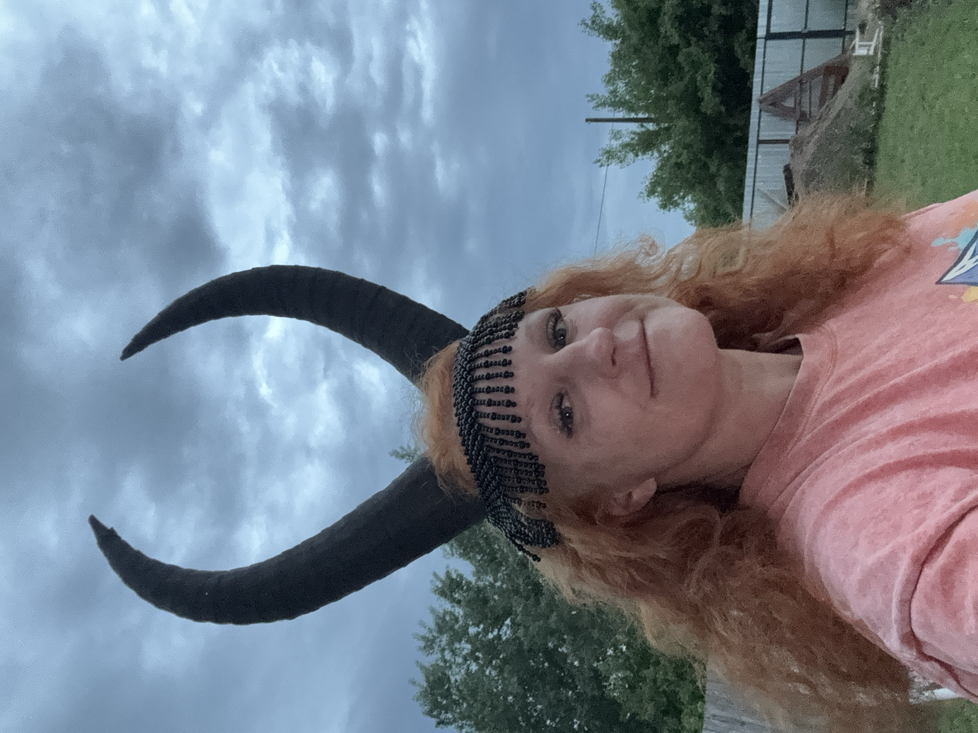 A woman with red hair is wearing a pink t-shirt, a beaded headpiece, and large black horns. The background includes a cloudy sky, trees, a fence, and what appears to be a pile of dirt or construction materials. The photo was taken in Chaikovsky, Russia.