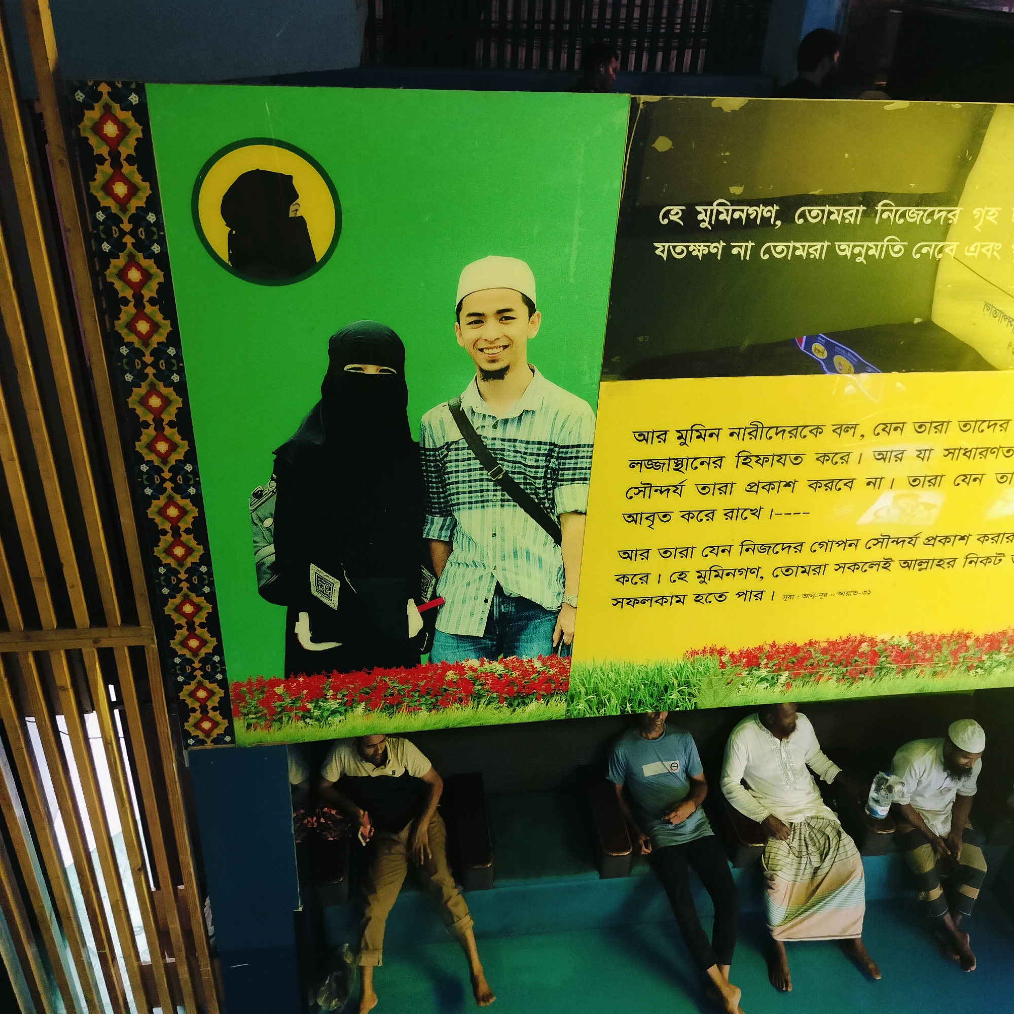 The image is taken in Dhaka, Bangladesh and shows a religious poster with the images of a man and a woman wearing Muslim attire. The woman is wearing a full face veil (niqab) and the man is wearing a taqiyah (Muslim cap). The poster also has text in Bengali script. Below the poster are four men sitting on a blue painted platform. They are barefoot and appear to be resting.