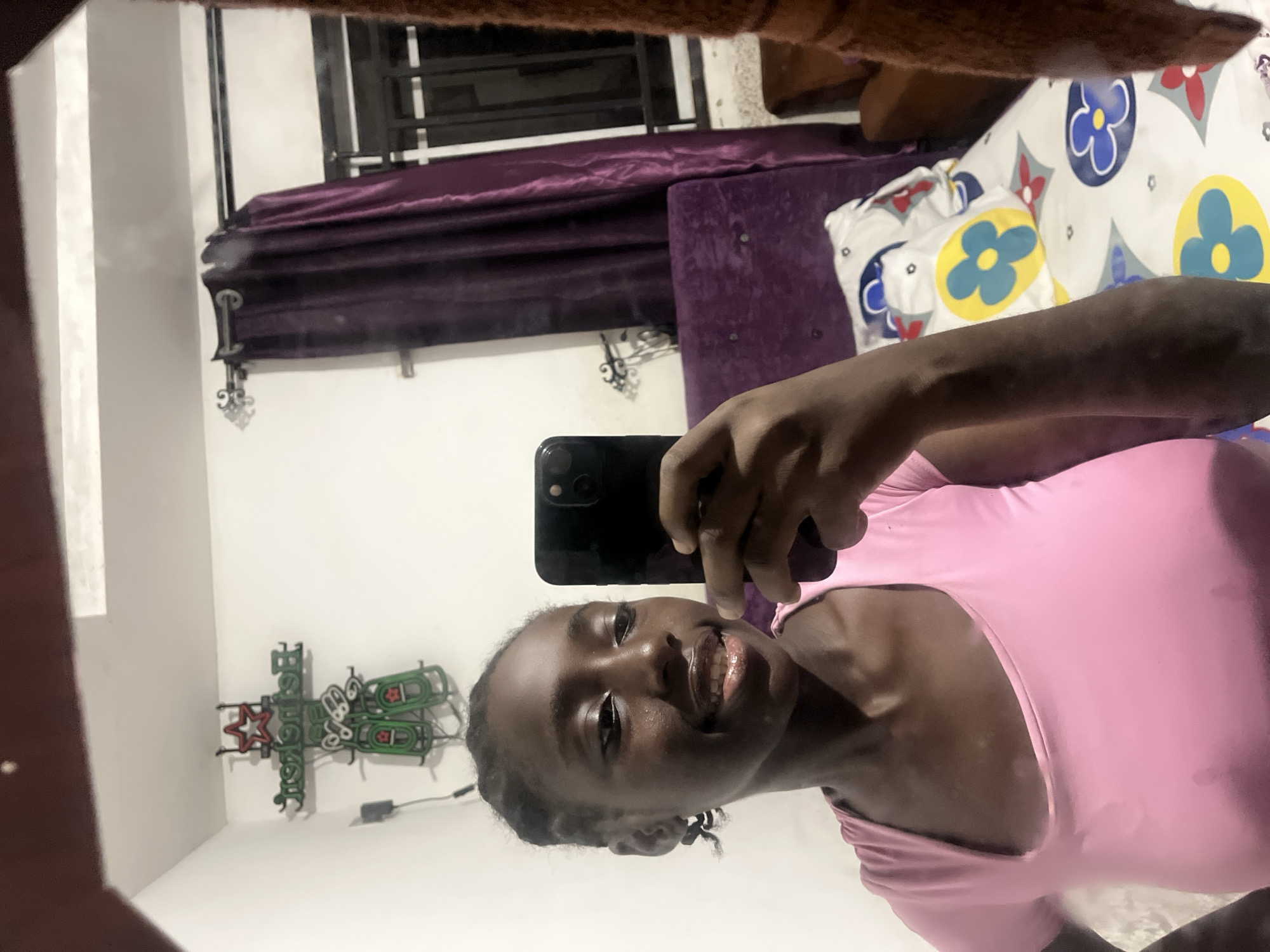 A selfie taken in a mirror by a young Black woman wearing a pink shirt. She is holding a black smartphone in her right hand. Behind her is a bed with purple velvet headboard and a white comforter with blue and yellow floral patterns. In the background, a window with a black frame can be seen. A wall decoration resembling a neon sign in green, red, and white colors is visible on the left. The location is most likely inside a home or apartment but there is not enough information to determine if the photo was taken in Abuja, Nigeria.