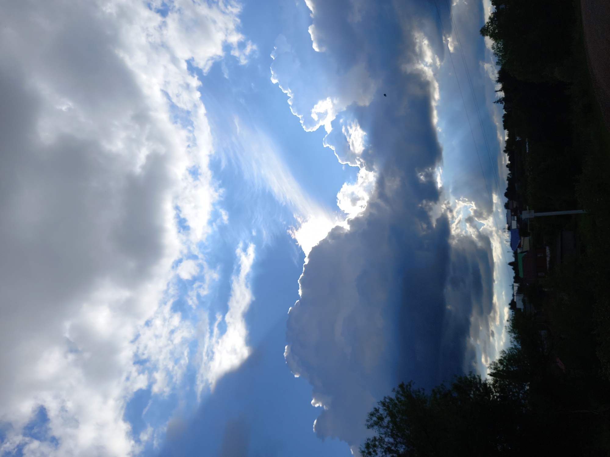 The image depicts a dramatic sky with a mix of light and dark clouds, suggesting a transition in weather. Sunlight breaks through the clouds, creating visible rays that illuminate the blue sky. The scene appears to be a rural or semi-rural setting, with hints of buildings and trees visible along the horizon, silhouetted against the bright sky. A single bird is visible as a small dark speck in the upper portion of the frame. The orientation of the image is vertical. The time of day is likely late afternoon or early evening, given the angle of the light and the dramatic cloud formations. The overall mood is one of awe and perhaps anticipation due to the powerful display of nature. There is no visible text in the image.