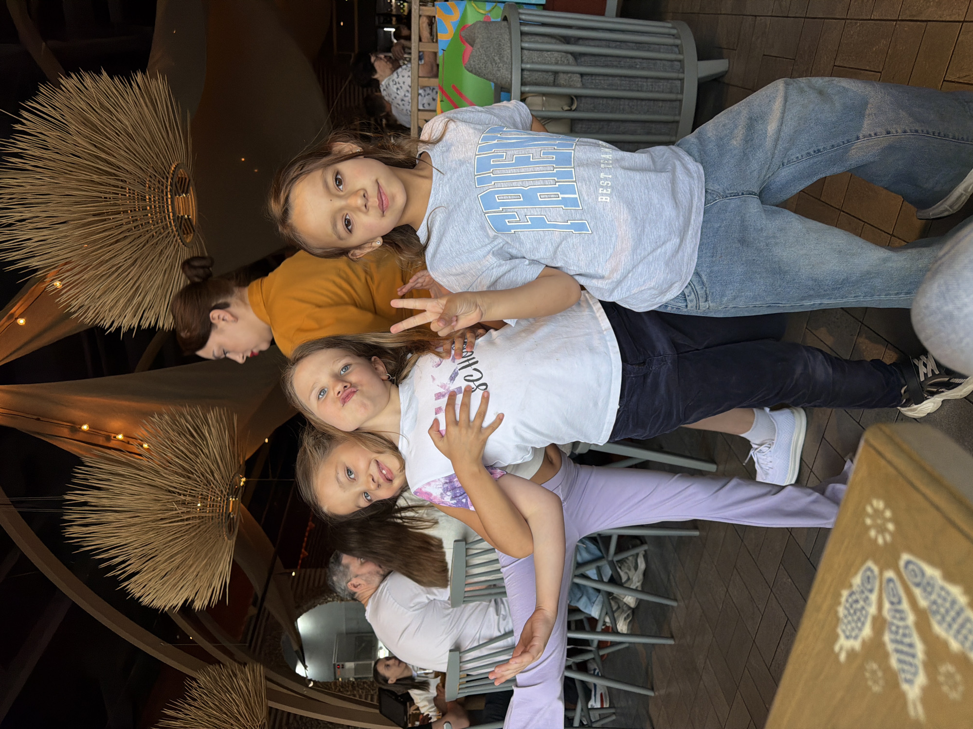 A group of people are in an indoor setting, likely a restaurant. Three young girls are prominently featured in the foreground. One wears a gray t-shirt and jeans, making a peace sign. A second girl wears a white t-shirt and black pants, and a third is in a white t-shirt with lavender pants. There are other individuals in the background including an adult woman in a yellow shirt, and an adult man in a white shirt. The setting includes decorative light fixtures resembling straw spheres. Chairs and tables are present. The location may be Krasnoyarsk, Russia.