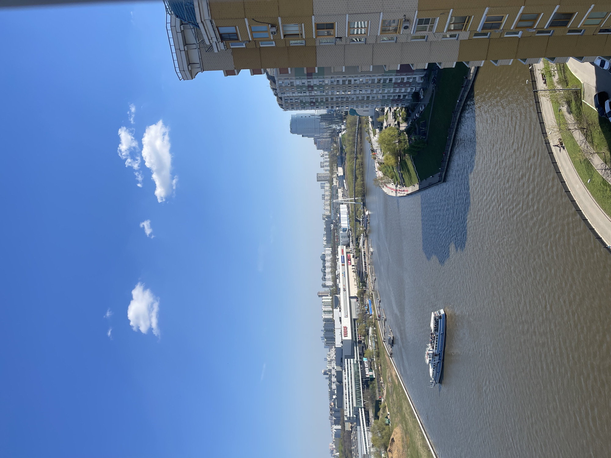 The image shows a view from a high vantage point overlooking a river in Moscow, Russia. The Moskva River winds through the city, with buildings and infrastructure lining its banks. A large passenger boat is seen navigating the river. The sky is blue with scattered white clouds. Buildings of varying heights and designs are visible, including residential apartments and commercial structures. A green parkway runs alongside the river.