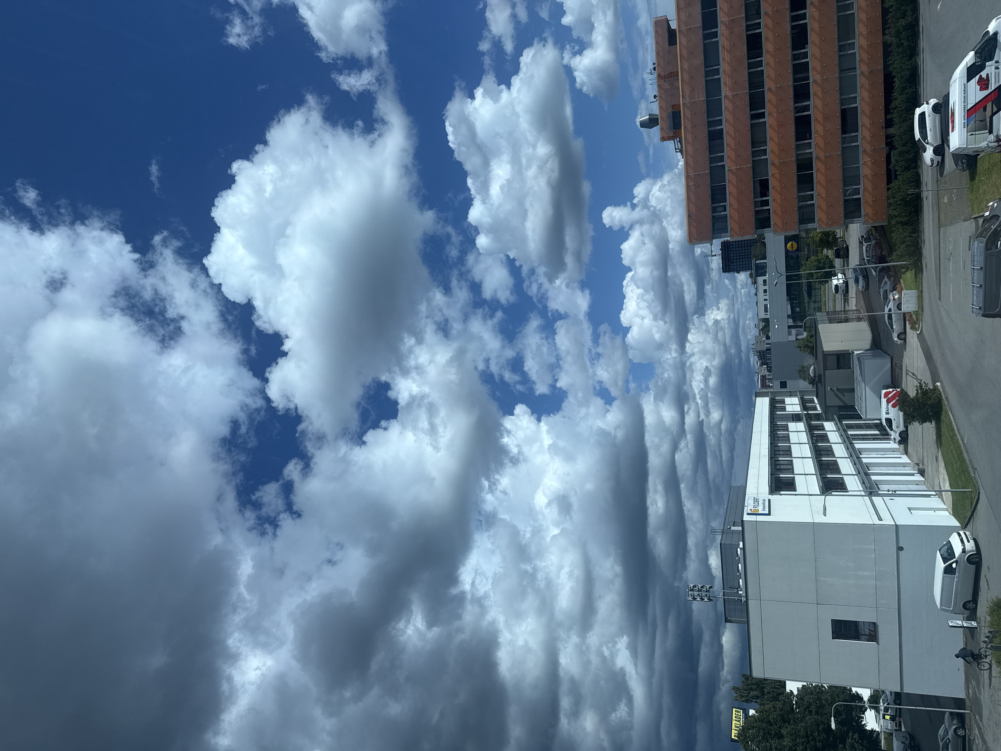 This is a photograph of a city scene in Tallinn, Estonia, under a partly cloudy sky. In the foreground, a white building with a modern design and large windows is visible on the left. To the right, a taller, orange-colored building with multiple floors and numerous windows dominates the right side of the image. Several vehicles, including white vans and cars, are parked in the lots and on the road. A person on a bicycle is seen on the left side of the image, near the white building. The sky above is a mix of bright blue patches and large, dramatic clouds, some of which are dark and appear to be gathering, suggesting a change in weather. The overall scene depicts an urban environment with a focus on commercial buildings and infrastructure.