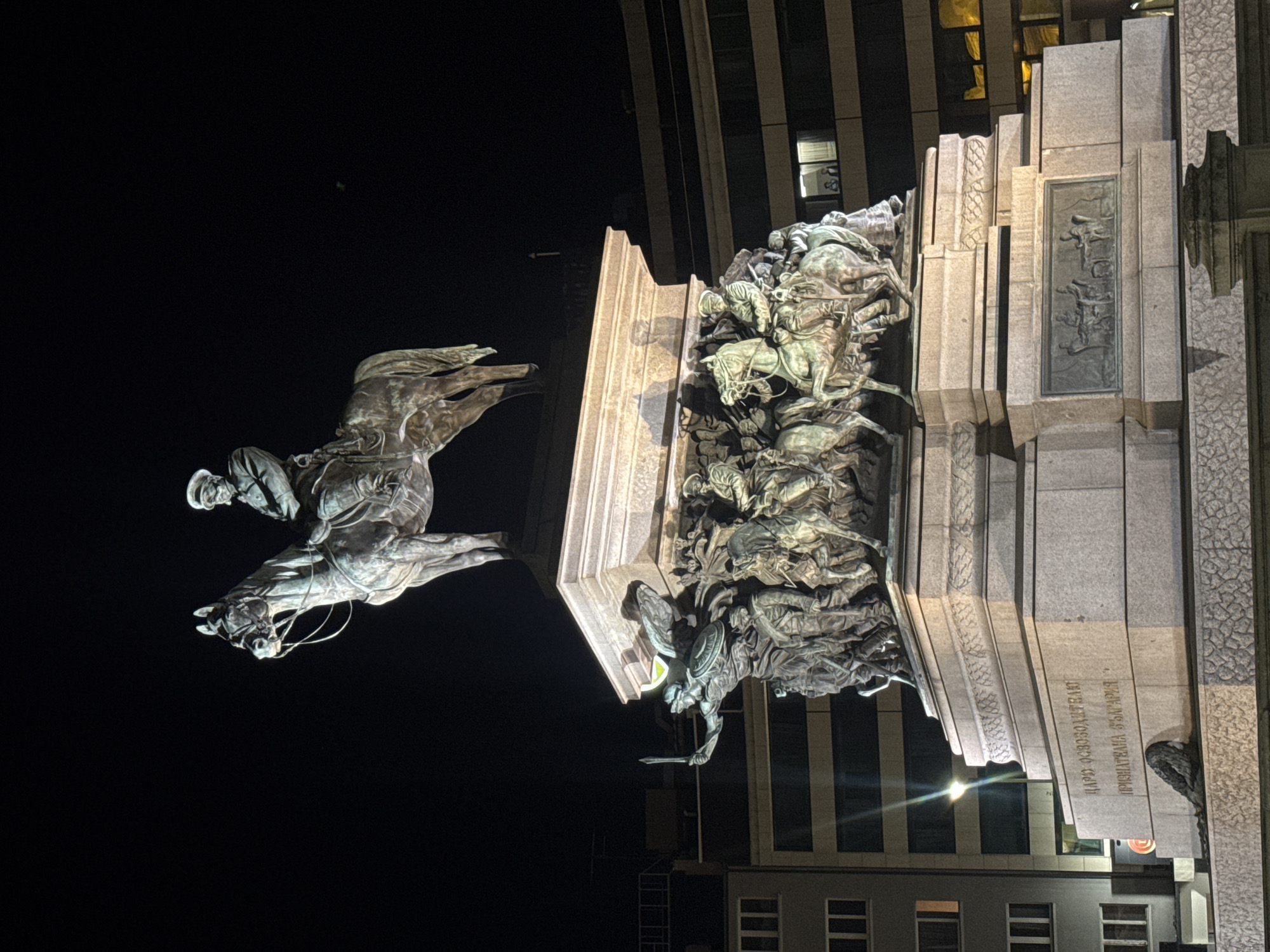 The image shows the Monument to the Liberators in Sofia, Bulgaria at night. The monument features a central equestrian statue of a figure wearing a military uniform and a cap, overlooking a group of bronze figures and horses at the base. The surrounding buildings are illuminated, and streetlights cast a glow on the monument. The inscription "ЦАРЬ ОСВОБОДИТЕЛЬ" and "ПРИЗНАТЕЛНА БЪЛГАРИЯ" can be seen on the monument's pedestal. The scene depicts a significant historical landmark in the city.