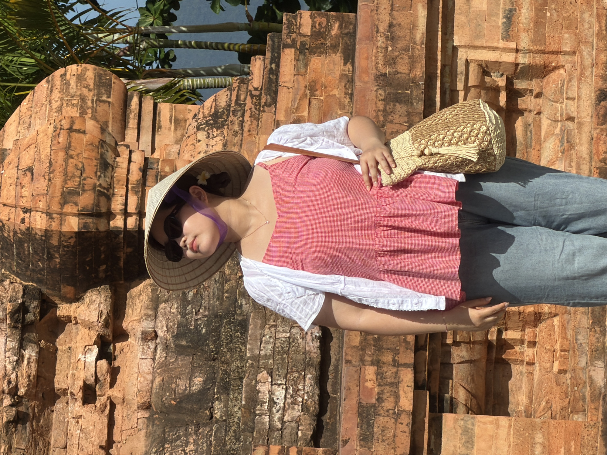A young woman stands in front of a brick structure, possibly a Cham tower, in Nha Trang, Vietnam. She wears a conical hat, sunglasses, a pink checkered peplum top, a white open cardigan, and blue jeans. A woven bag with a tassel hangs from her shoulder. Palm trees and a blue sky are visible in the background. The brickwork is aged and weathered, with patches of moss and discoloration. The lighting suggests it is daytime, with strong sunlight casting shadows.