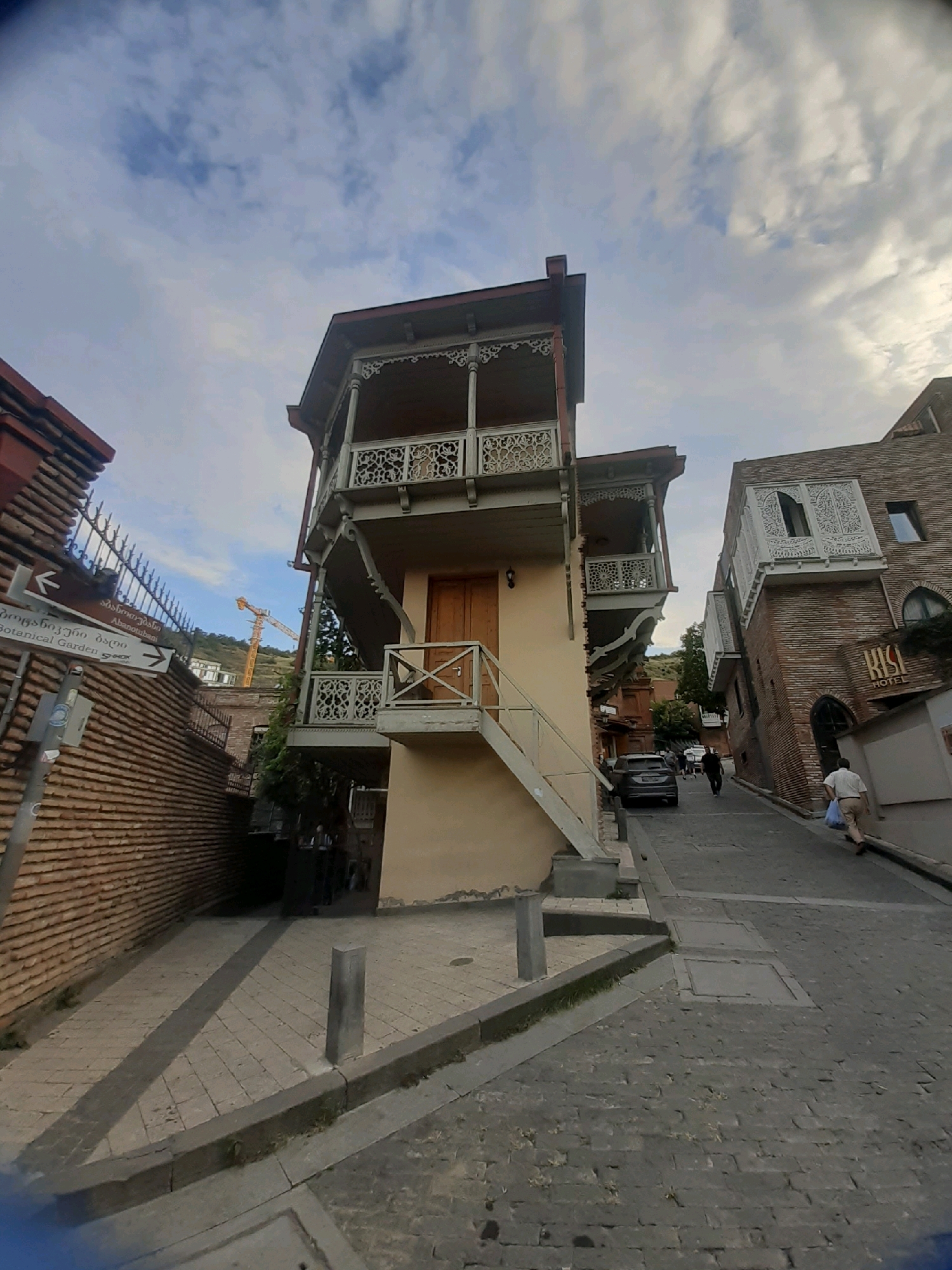 The image depicts a street scene in the old city of Tbilisi, Georgia, characterized by traditional architecture. In the foreground, a narrow cobblestone street slopes uphill. On the left, a brick wall with a metal fence borders a pedestrian area paved with light-colored tiles. A street sign points left, with inscriptions in Georgian and English indicating "Abanotubani" and "Botanical Garden". Dominating the center and right of the frame are two multi-story buildings. The building on the left is a narrow, light-yellow structure with a prominent wooden balcony featuring intricate carvings and a steep staircase leading to its entrance. The building on the right is a brick structure with decorative white balconies and arched windows, a sign for "RUSTAVELI HOTEL" is visible on its facade. In the background, a yellow construction crane is visible against a cloudy sky. Several individuals are present: one person is walking up the street on the right, carrying a light blue bag, and another person is walking away from the camera further up the street. A dark SUV is parked on the left side of the street. The overall atmosphere suggests a historic urban environment.