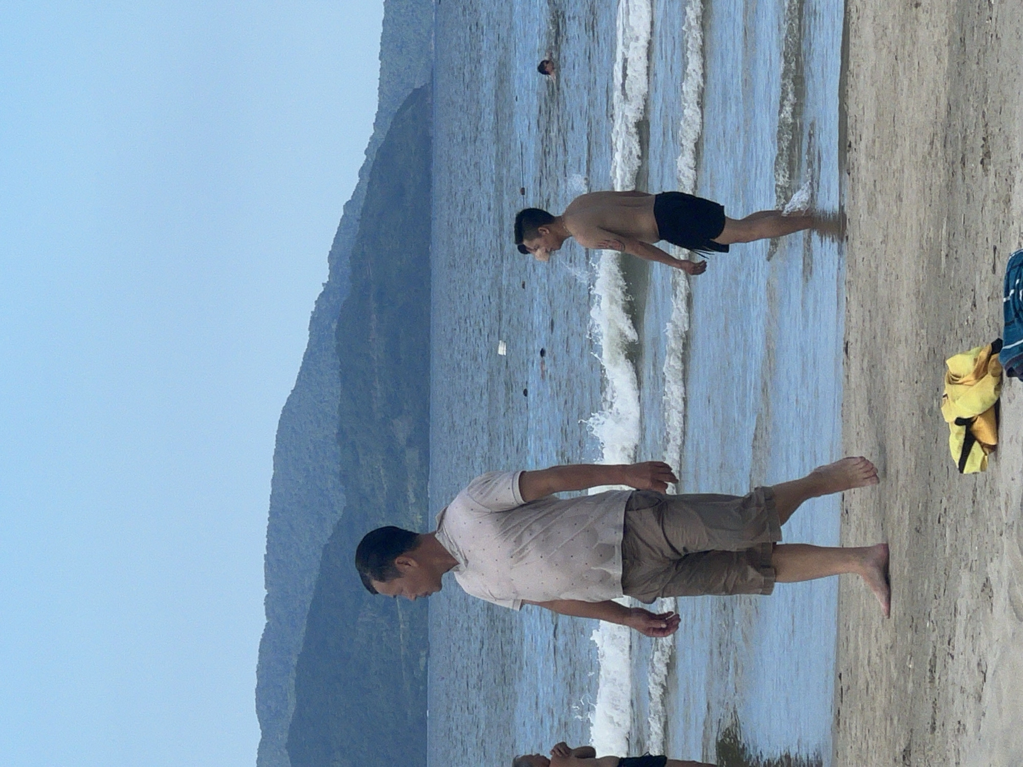 The image depicts a beach scene at My Khe Beach in Da Nang, Vietnam. In the foreground, a man wearing a light-colored short-sleeved shirt with a pattern and brown shorts walks barefoot on the sand. Further back, a man in black swim shorts stands in the shallow water, with waves breaking around his legs. Several other people are visible in the ocean, some swimming and others further out. The background features rolling green hills covered in vegetation, typical of the coastal landscape surrounding Da Nang. The sky is clear and pale blue. On the sand in the foreground, there are some folded yellow and blue towels. The overall impression is of a relaxed, casual day at the beach.