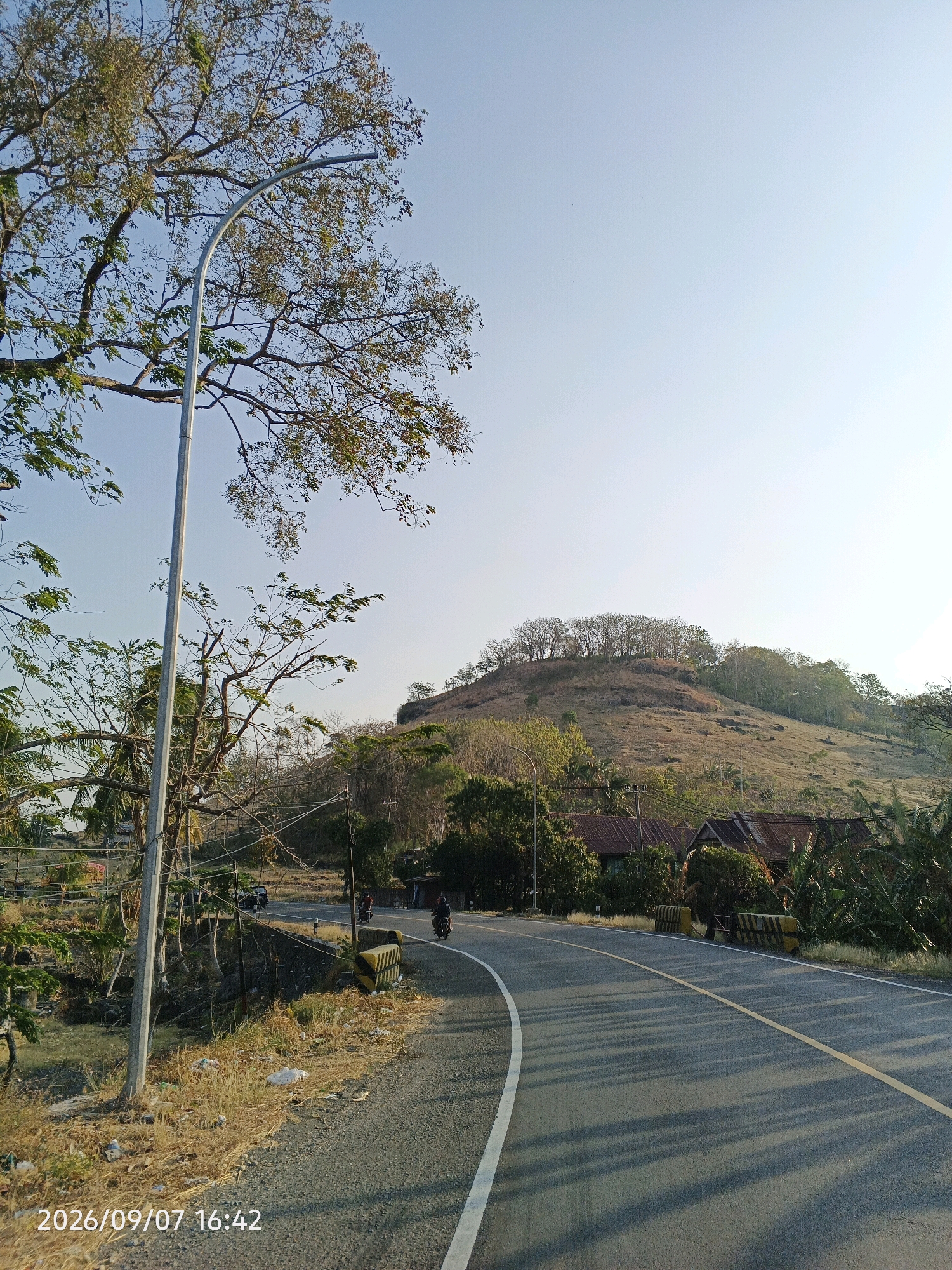 This image depicts a rural road in Lainungan, Indonesia, on September 7, 2026, at 4:42 PM. The road curves to the right, with a yellow center line and a white edge line. A lamppost stands on the left side of the road, with a large tree casting shadows. In the background, a dry, grassy hill is topped with a cluster of trees. Several houses with corrugated roofs are visible nestled at the base of the hill, surrounded by vegetation. Two motorcycles are seen on the road, one traveling away from the viewer and another appearing to be parked. The sky is clear and blue. The ground along the roadside is covered in dry grass and scattered litter.