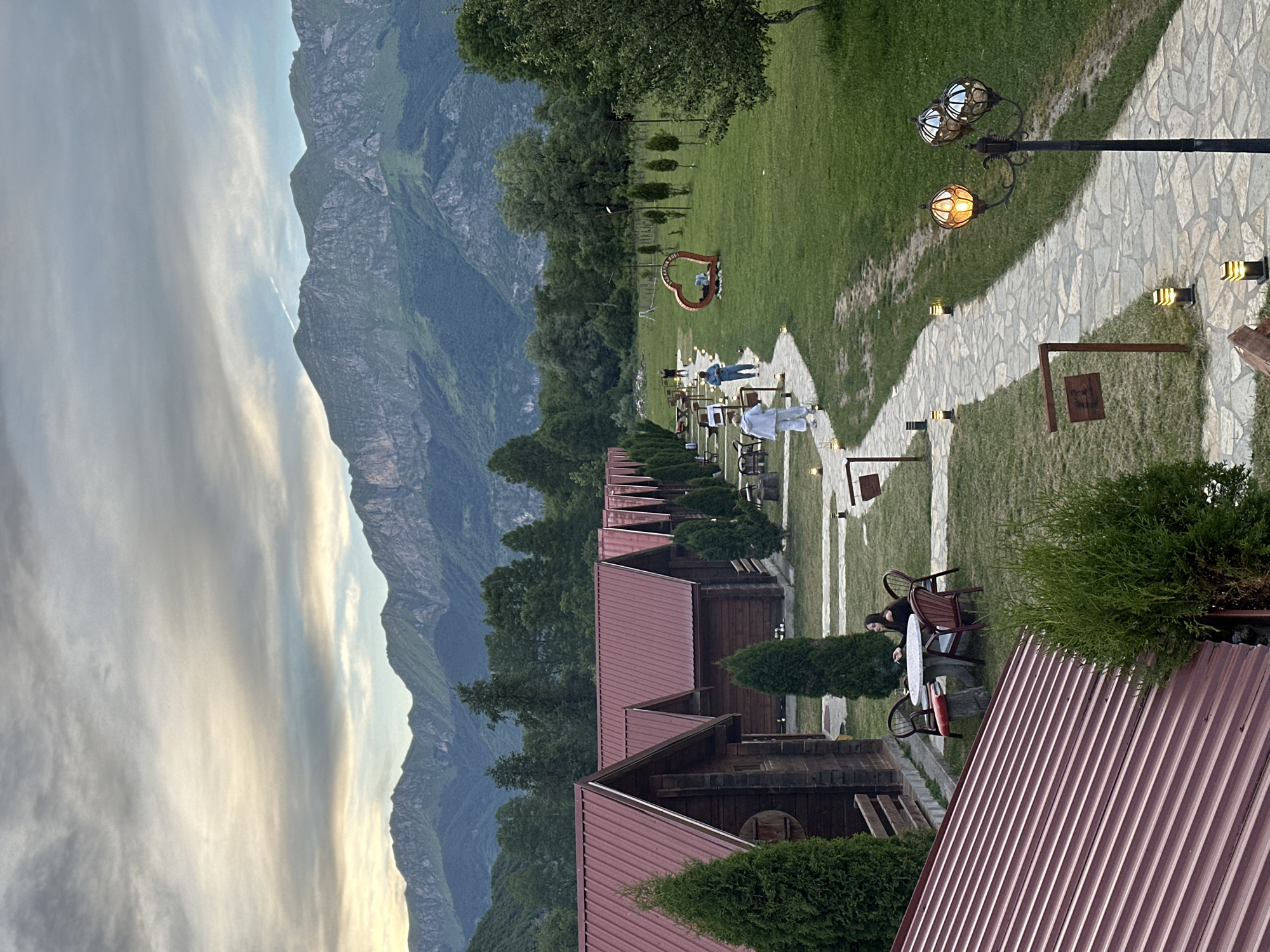 The image depicts a serene resort nestled in Gusinje, Montenegro, with a majestic mountain range serving as a dramatic backdrop. The foreground is dominated by a row of charming wooden cabins, each topped with a vibrant red metal roof. A paved pathway winds its way through the meticulously manicured lawn, illuminated by decorative lampposts and smaller ground lights.
Several individuals are visible in the scene. Two people, dressed in casual attire, are strolling along the pathway. Another person, seated at a round table outside one of the cabins, appears to be enjoying a cigarette. Further down the path, a heart-shaped sculpture stands prominently on the green expanse, with a person relaxing within its embrace.
The environment is lush and verdant, with trees and manicured bushes dotting the landscape. The sky above is a canvas of soft clouds, hinting at either the fading light of dusk or the gentle break of dawn. The overall impression is one of tranquility and natural beauty, characteristic of a peaceful mountain retreat.