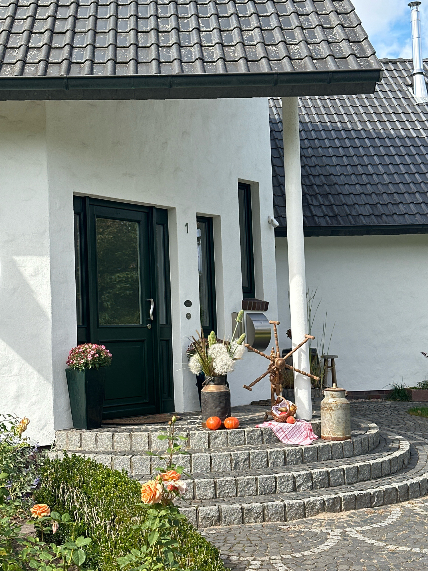 The image depicts the entrance of a modern white house located in the Samtgemeinde Thedinghausen, Germany. The house features a dark grey tiled roof and a dark green front door with glass panels. The entrance is accessed by a curved set of granite steps, with a cobblestone pathway leading away from them.
The scene is decorated for autumn. On the top step, two small orange pumpkins are placed, accompanied by a basket containing more pumpkins and a red and white checkered cloth. To the left of the pumpkins is a dark gray milk can. To the right of the pumpkins, a large dark gray vase holds dried grasses and what appears to be dandelions. A rustic, wooden spinning wheel-like object is positioned behind the vase and to the left of a small wooden stool.
To the left of the front door, a dark green planter holds a cluster of pink flowers. In the foreground, a lush green hedge is visible, with several peach-colored roses blooming. The overall impression is one of a well-maintained residential property with seasonal decorations. There are no people or animals present in the image.