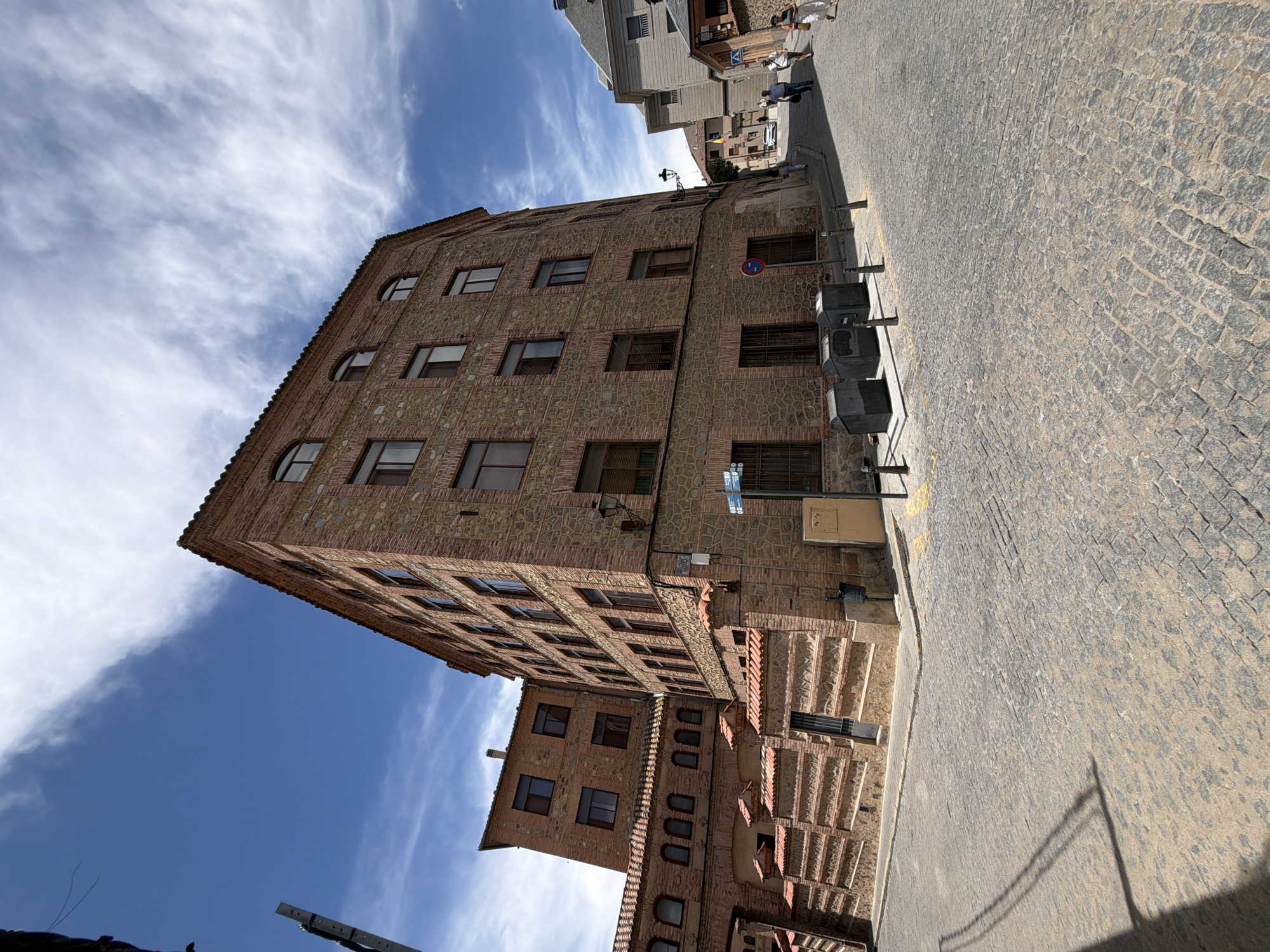 The image depicts a cobblestone street in Segovia, Spain, featuring historical architecture. The primary subject is a multi-story brick building with stone accents and arched windows, characteristic of European old towns. The building's facade is textured with a mix of red brick and rough-hewn stone. Adjacent to it, another section of the building features a lower brick wall with a terracotta tiled roof and rounded archways.
The street itself is paved with irregularly shaped cobblestones, worn smooth by time and traffic. The scene is set under a bright blue sky with scattered white clouds, suggesting fair weather. In the background, more buildings with similar architectural styles are visible, lining the ascending street.
Several people are present on the street. In the middle ground, two individuals are walking away from the camera, dressed in casual attire. Further up the street, a group of three people, two women and a man, are also walking away from the camera, with one woman in the foreground wearing a white dress and carrying a bag. On the right side of the street, a signpost is visible near a building.
Essential objects include trash receptacles located near the building's entrance and a lamppost. The overall impression is one of a charming, historic urban environment in Segovia, Spain, likely a tourist area.