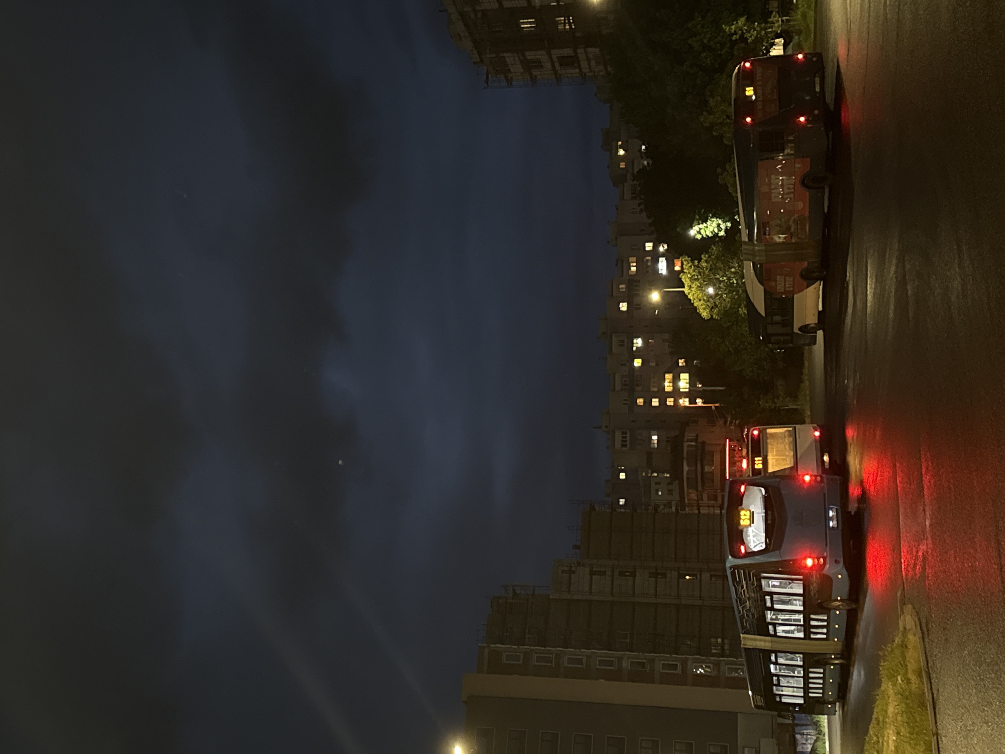 The image depicts a night scene in Burgas, Bulgaria, likely on a street lined with tall apartment buildings. The sky is dark with clouds. Two buses are parked on the right side of the frame, with their rear lights illuminated. The buildings across the street have lit windows. The road surface appears wet, reflecting the lights. No people are visible in the image.