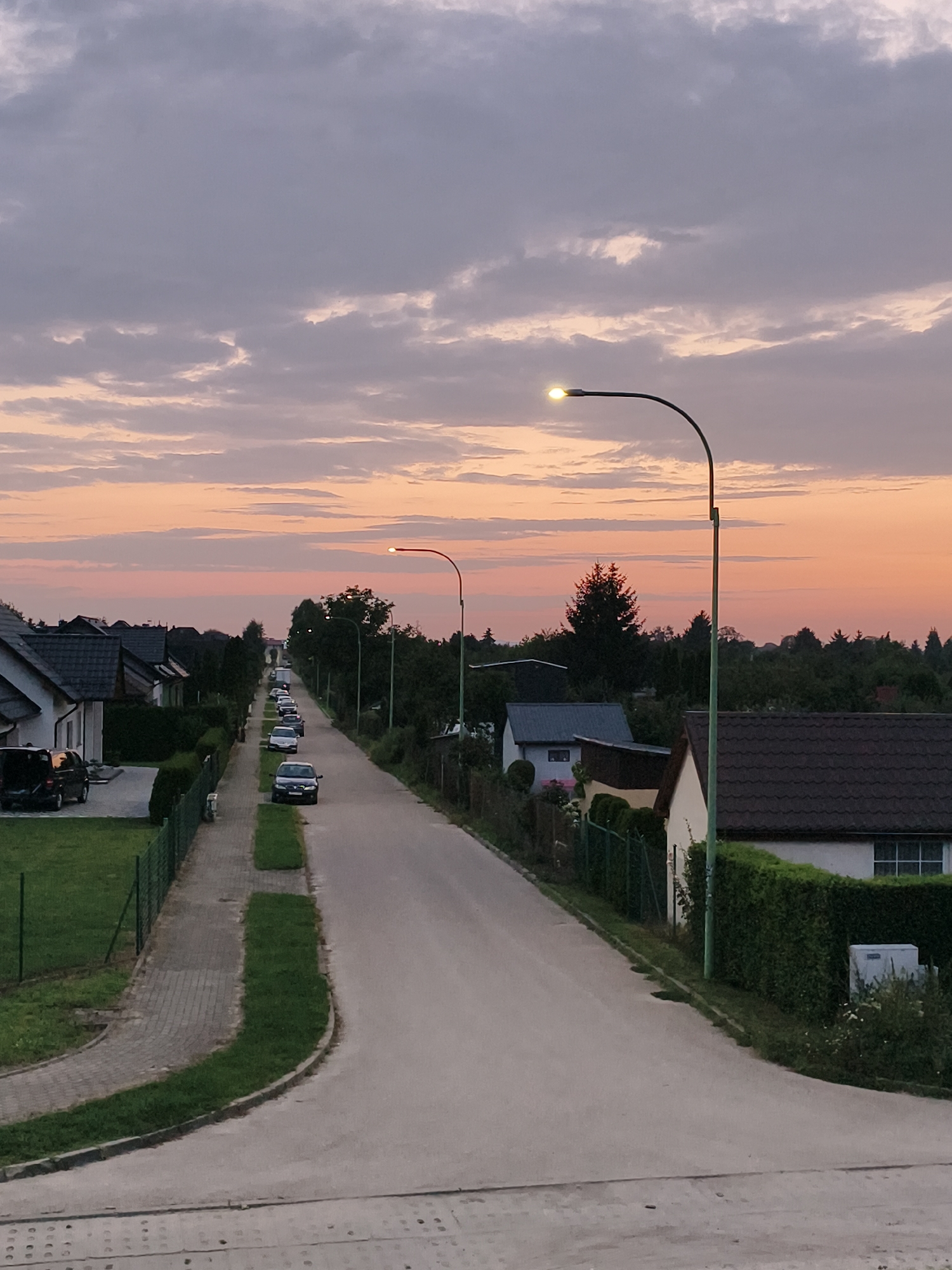 The image depicts a quiet suburban street in Tczew, Poland, at dusk. The sky is a gradient of soft pinks and grays, with scattered clouds. A street lamp illuminates the scene, casting a warm glow on the asphalt road that curves gently uphill. Several cars are parked along the street, and houses with dark tiled roofs line both sides. Green hedges and manicured lawns contribute to the orderly appearance of the neighborhood. The overall atmosphere is peaceful and serene, suggesting the end of a day in this Polish town.