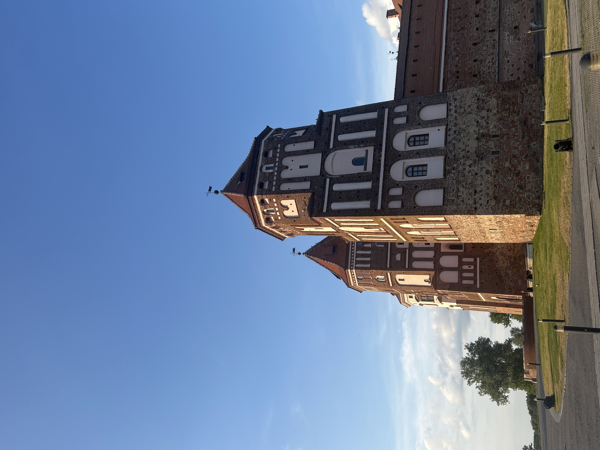The image shows a view of Mir Castle, a UNESCO World Heritage site located in the settlement of Mir in the Karelichy District of Grodno Region, Belarus. The castle is a large, multi-towered fortress constructed from red brick and stone. The sky above is a clear blue with a few wispy clouds. In the foreground, a paved area leads up to a grassy slope that surrounds the castle walls. There are several decorative lampposts and a historical cannon positioned on the grounds. Trees are visible in the background.