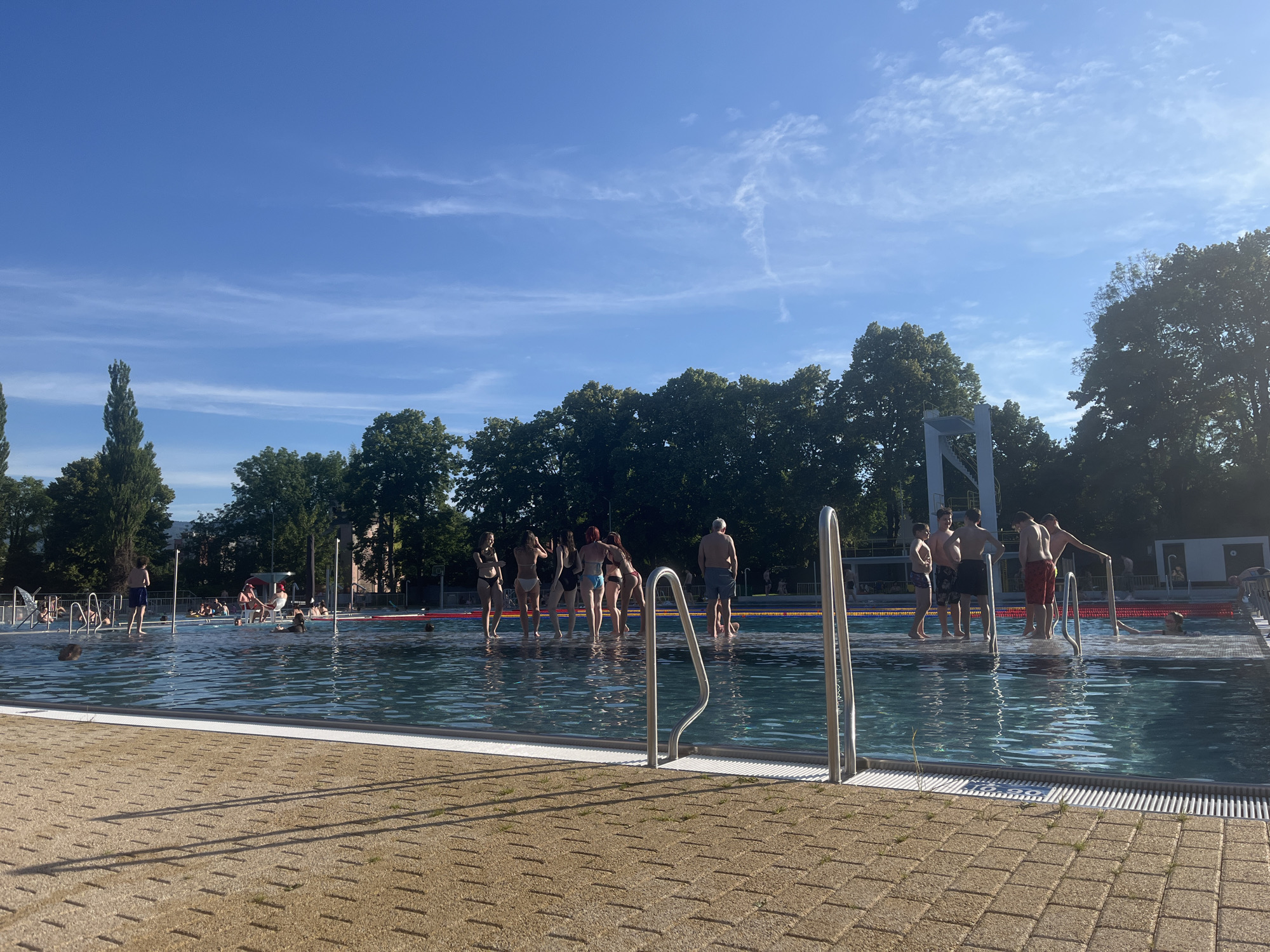 In an outdoor swimming pool, a group of people are gathered. Some are standing in the shallow end of the pool, while others are on the pool deck. The sun is shining brightly, casting long shadows on the ground. The water is clear and blue, and there are trees and buildings in the background. The image captures a recreational scene on a sunny day.