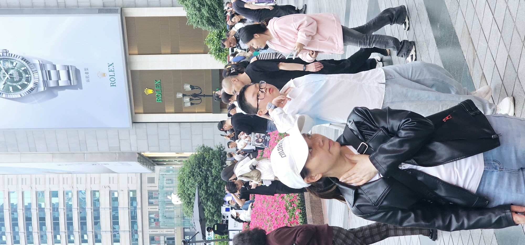 The photograph captures a scene on a paved area in Yuzhong District, China. People are lined up, and appear to be taking pictures or waiting. In the background, a Rolex store is visible with an advertisement of a watch. The environment suggests a commercial street with buildings. The crowd consists of a mix of ages, some wearing casual clothing like jeans and jackets. Flowers are visible at the edge of the waiting area.