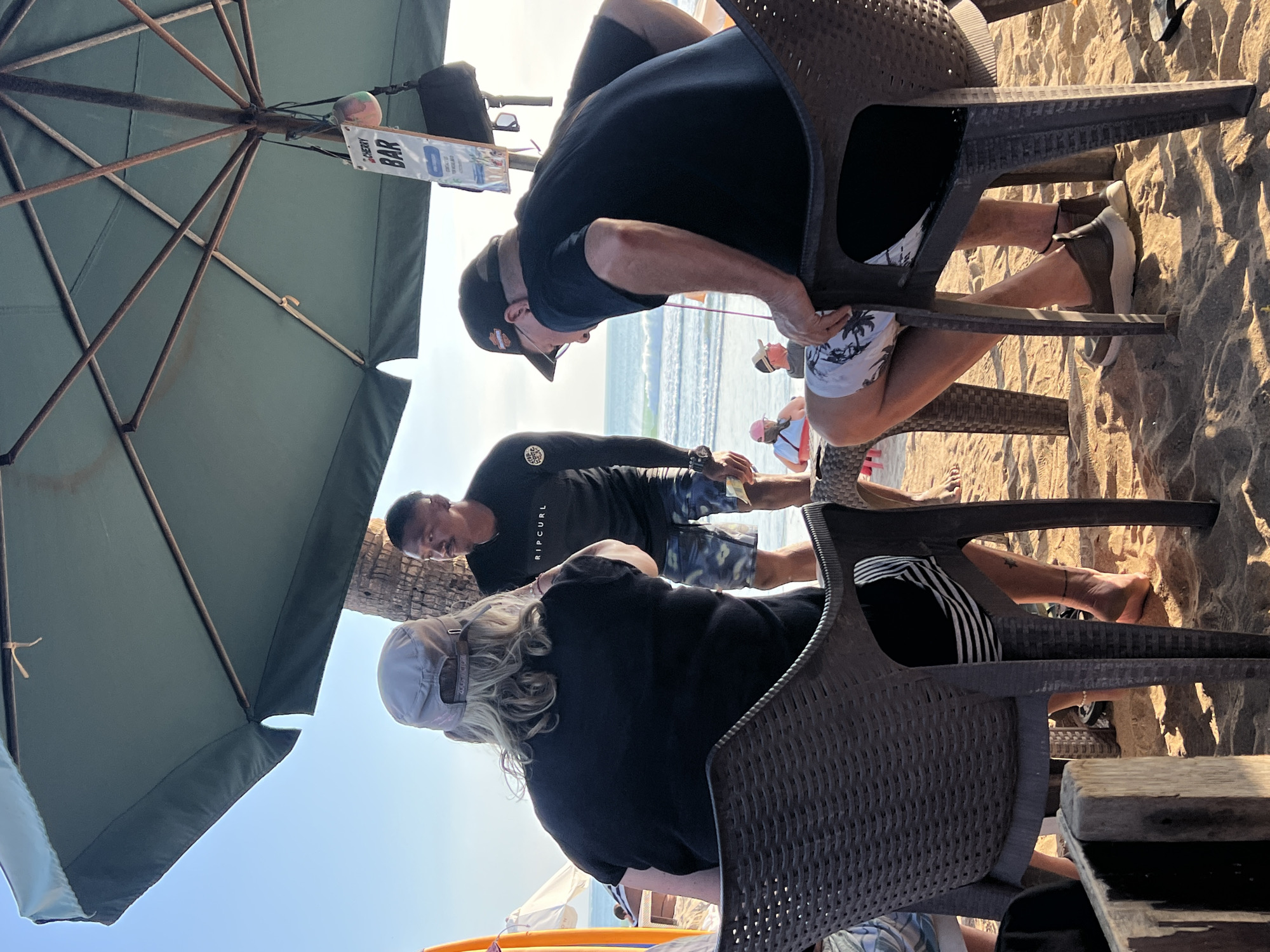 At a beach bar in Kuta, Indonesia, three individuals are present. One person, viewed from behind, wears a black t-shirt and a light-colored baseball cap, with blonde hair visible. Another person, facing the first, is dressed in a black long-sleeved shirt with "RIPCURL" printed on it, and patterned shorts. The third individual, also viewed from behind, wears a black t-shirt and a baseball cap. They are seated on dark-colored plastic chairs on the sandy beach. An umbrella provides shade, and a sign for "CHERRY BAR" hangs from it. The ocean and waves are visible in the background, with a surfboard standing upright to the left. Several other people are in the background, some seated and some standing, and a beach umbrella is visible. The lighting suggests it is daytime.