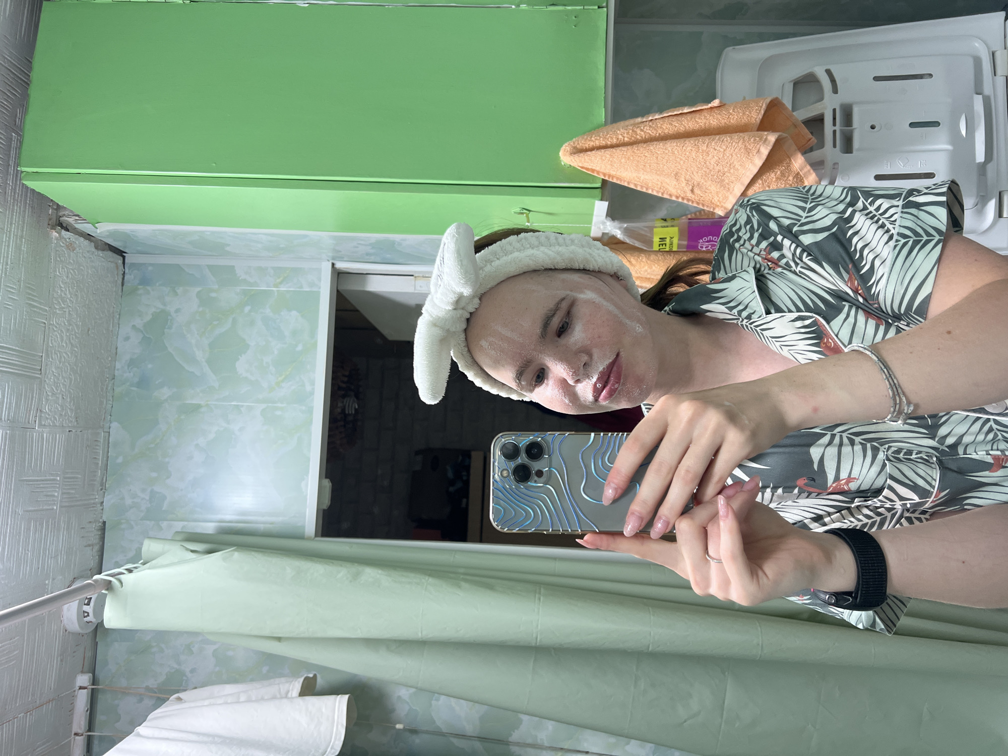 A young woman with a white headband and a face mask is taking a selfie in a bathroom. She is wearing a patterned pajama top. The bathroom walls are tiled with a light green marble pattern. A green cabinet is visible on the right side of the image, and an orange towel hangs from it. A white shower curtain is on the left. The ceiling appears to be made of textured panels. The woman is holding a smartphone with a decorative case.