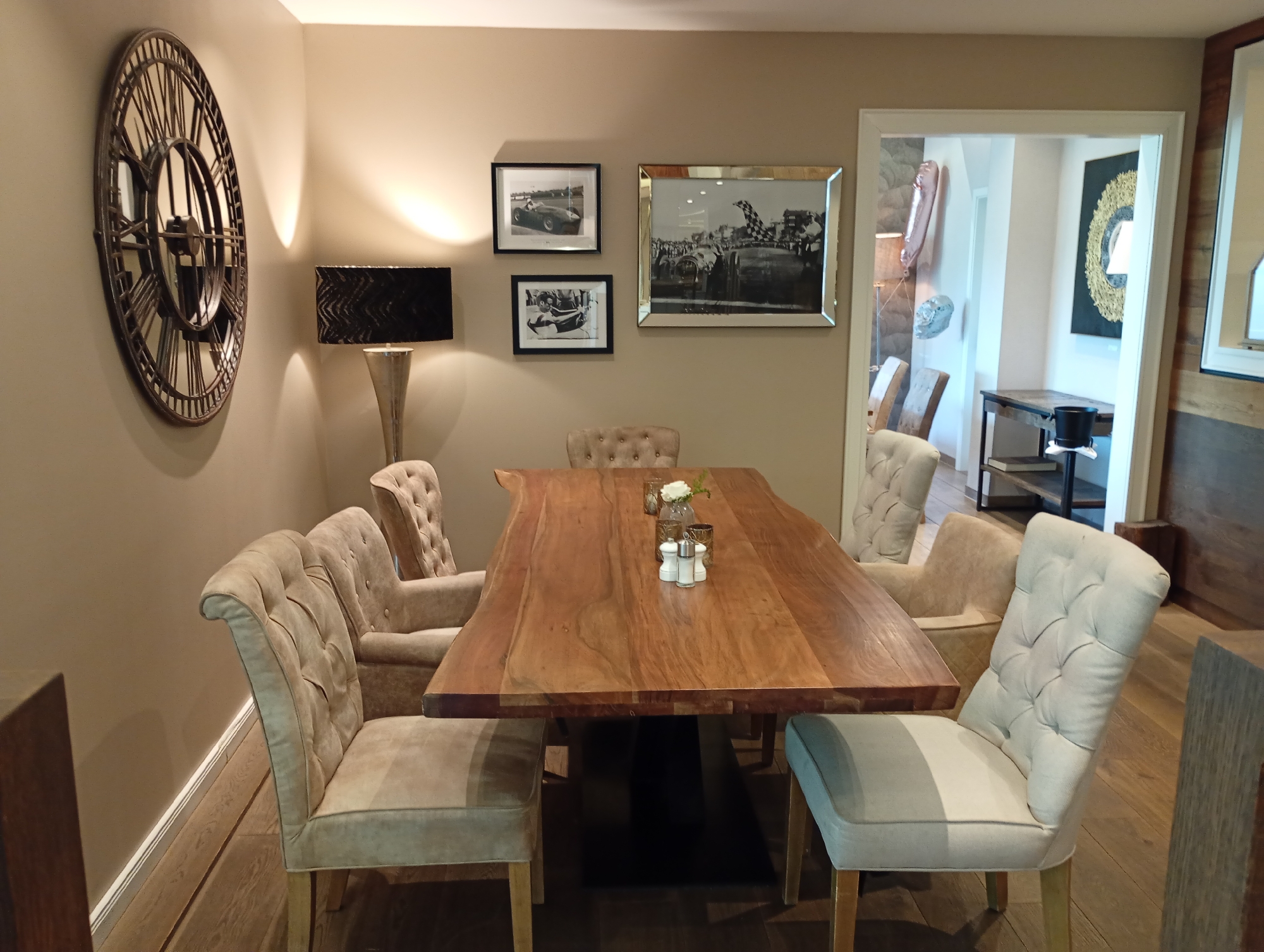 The image depicts a dining area with a large wooden table surrounded by tufted upholstered chairs. The room is neutrally decorated with light-colored walls. On the wall to the left, a large round clock with Roman numerals is prominently displayed. To the right of the clock, a table lamp with a textured shade sits on a stand. Several framed black and white photographs are hung on the wall, featuring images of vintage cars. A large, ornate framed photograph of a car race is also visible. The chairs around the table are upholstered in a neutral fabric and feature button tufting. A small vase with a white flower and a salt and pepper shaker set are placed on the table. The floor is made of wooden planks. Through an open doorway on the right side of the image, a glimpse of another room is visible, showing a console table with shelves and a black pot. The overall ambiance suggests a stylish and comfortable dining space. This specific room is located in the "Romantik Hotel Sachsen-Anhalt" in Bad Sachsa, Germany.