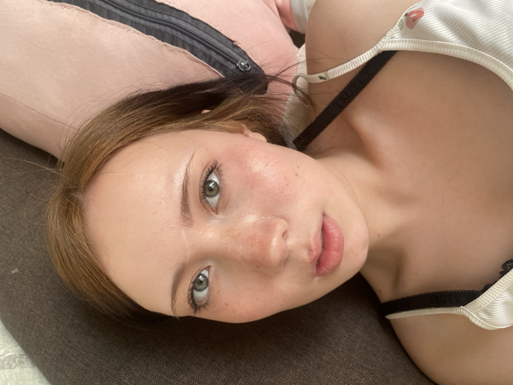 The image shows a young woman lying down, possibly on a couch or bed, with a pink pillow visible behind her head. She is wearing a white top with a black lace strap. Her skin is fair and shows some freckles. She has light brown hair and green eyes, and is looking directly at the camera with a neutral expression. The background is a dark brown textured surface.
The location is in the city of Unknown, Unknown.