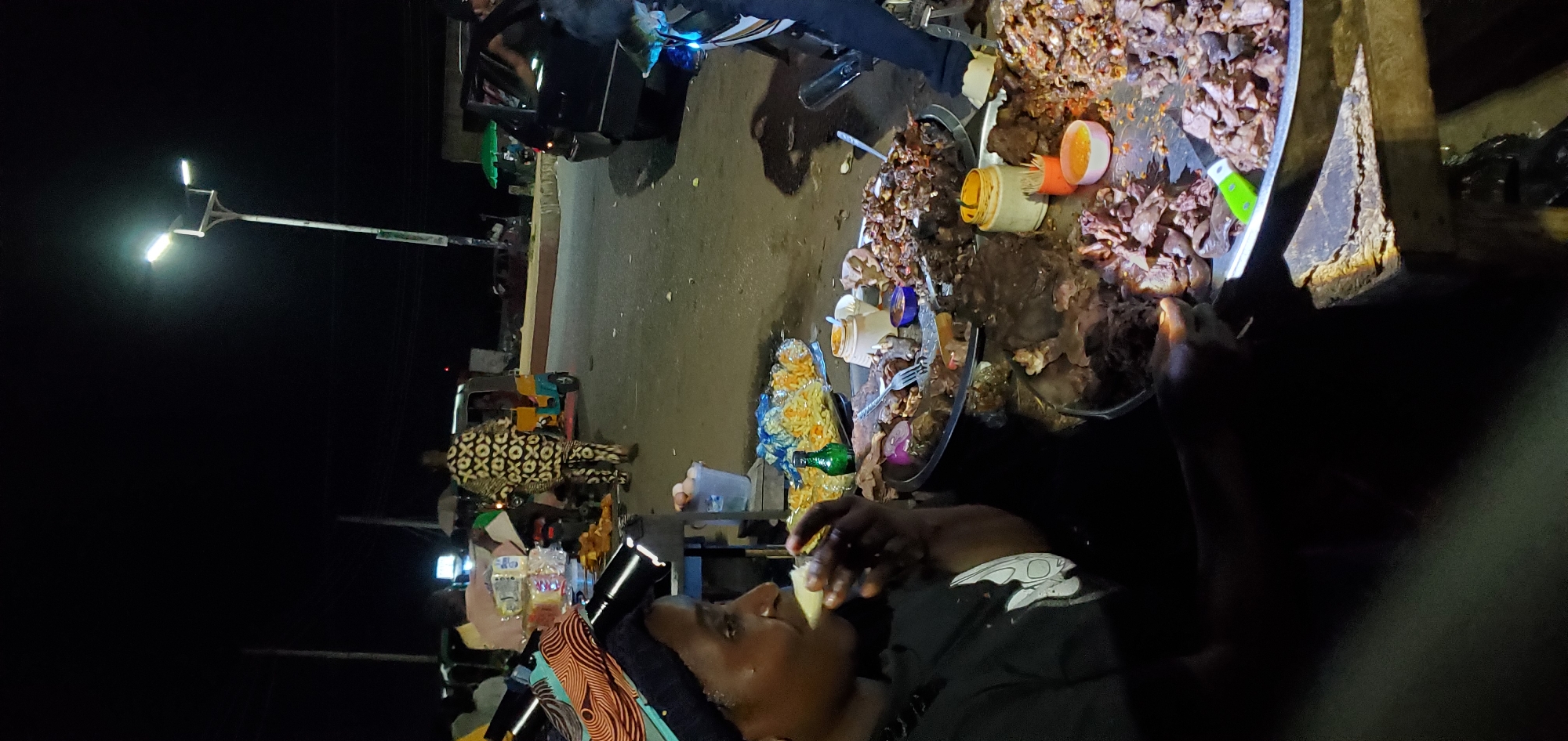 The image depicts a street scene at night in an unidentified location. A man is shown eating something, while behind him is a table or surface laden with cooked meat and various condiments in containers. Other items, including what appears to be bottles and snacks, are also present. A street light illuminates part of the scene. Further back, a motorized vehicle (possibly a tuk-tuk) and another individual wearing a patterned outfit are visible. The environment appears to be an urban or semi-urban street setting.