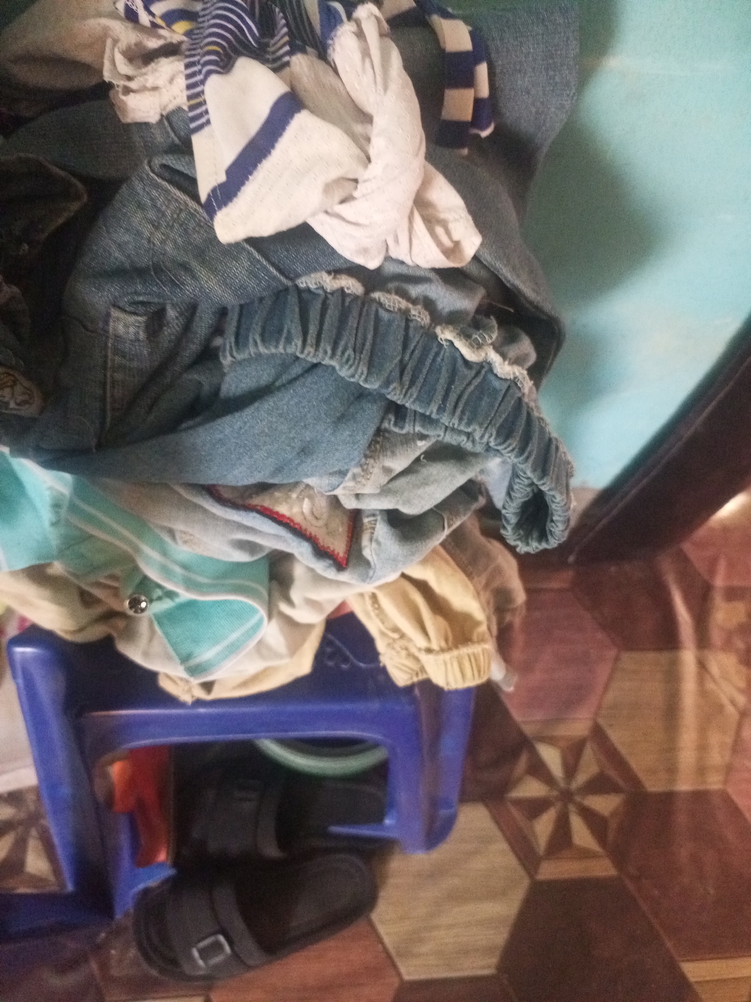 The image displays a pile of laundry and a blue plastic stool on a floor with hexagonal tiling. The laundry consists of various garments, including jeans, t-shirts, and striped fabric. A pair of black sandals is visible beneath the stool. The background includes a light blue surface and a dark brown object. The scene appears to be indoors, possibly in a residential setting in Jalingo, Nigeria.
