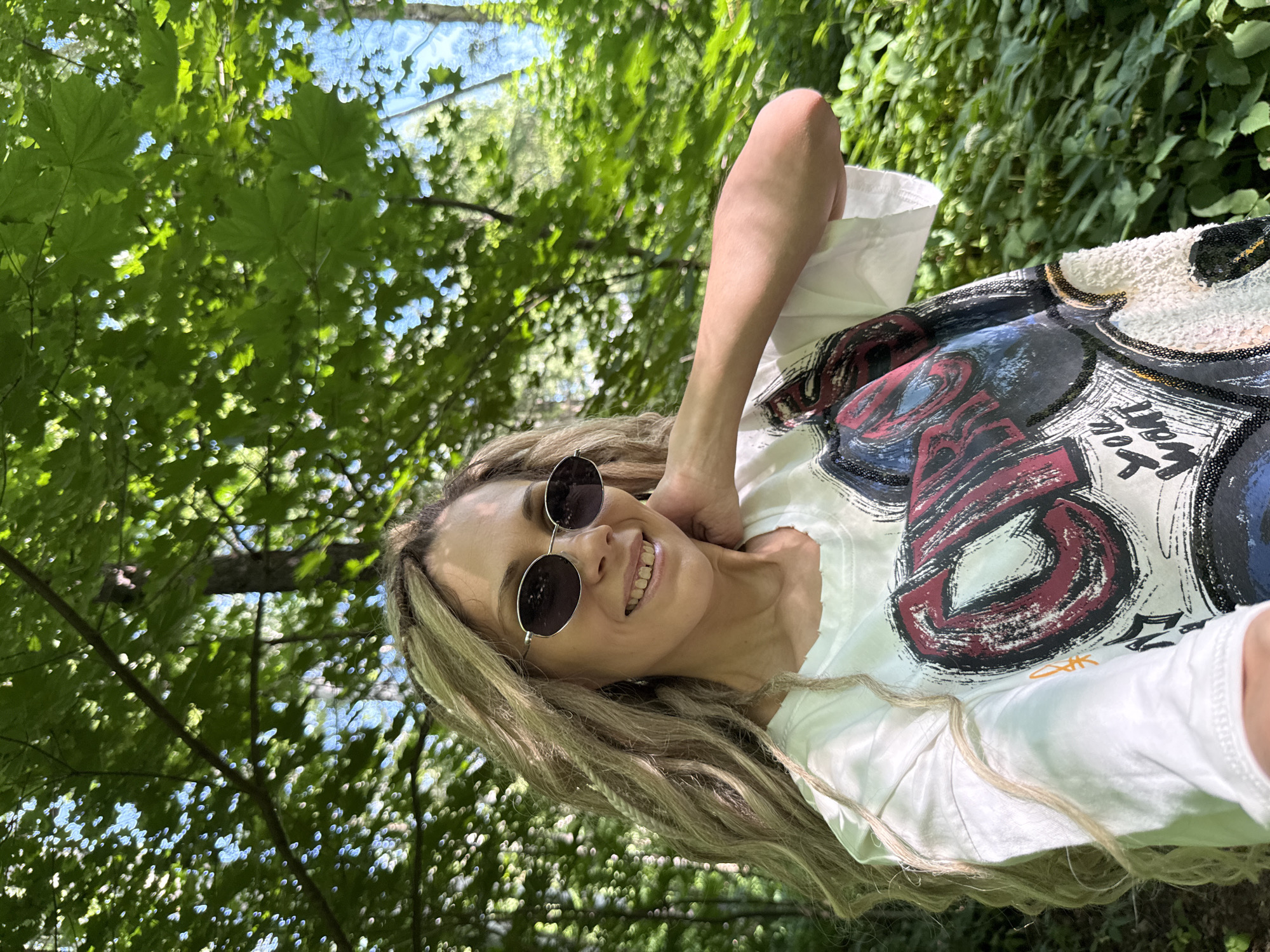 A woman with blonde dreadlocks and sunglasses smiles while posing for a selfie outdoors. She is wearing a white t-shirt with a graphic print. The background is a forest with lush green trees and dappled sunlight. The location is Rusanovo, Russia.