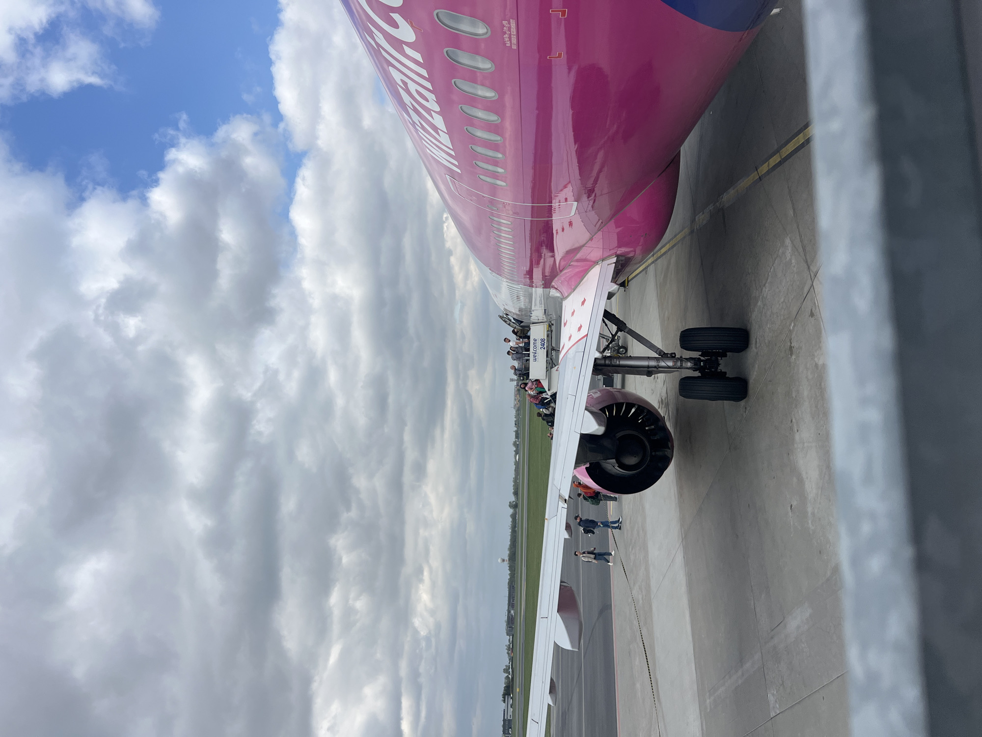 The image displays a magenta-pink Wizz Air aircraft, likely an Airbus A320 family plane, parked on an airport tarmac during the daytime. Passengers are actively boarding the aircraft via a mobile stairway positioned at the front left door. Several individuals, dressed in casual attire, are visible on the stairway and near the aircraft's entrance.
The airplane's fuselage features the prominent "Wizzair" logo in white letters, alongside smaller Arabic script visible above some of the windows. The mobile boarding stairway bears the text "WELCOME 2408" on its side. One of the aircraft's jet engines is visible beneath the wing, along with parts of the landing gear.
The sky is partly cloudy to overcast, with white and grey clouds dominating, though some patches of blue sky are also visible. The ground is a dark grey tarmac with yellow painted lines. In the background, beyond the wing and tarmac, a grassy field extends, suggesting the surroundings of an airfield, consistent with an airport in Warsaw, Poland. The scene captures a typical moment of passenger boarding before a flight.