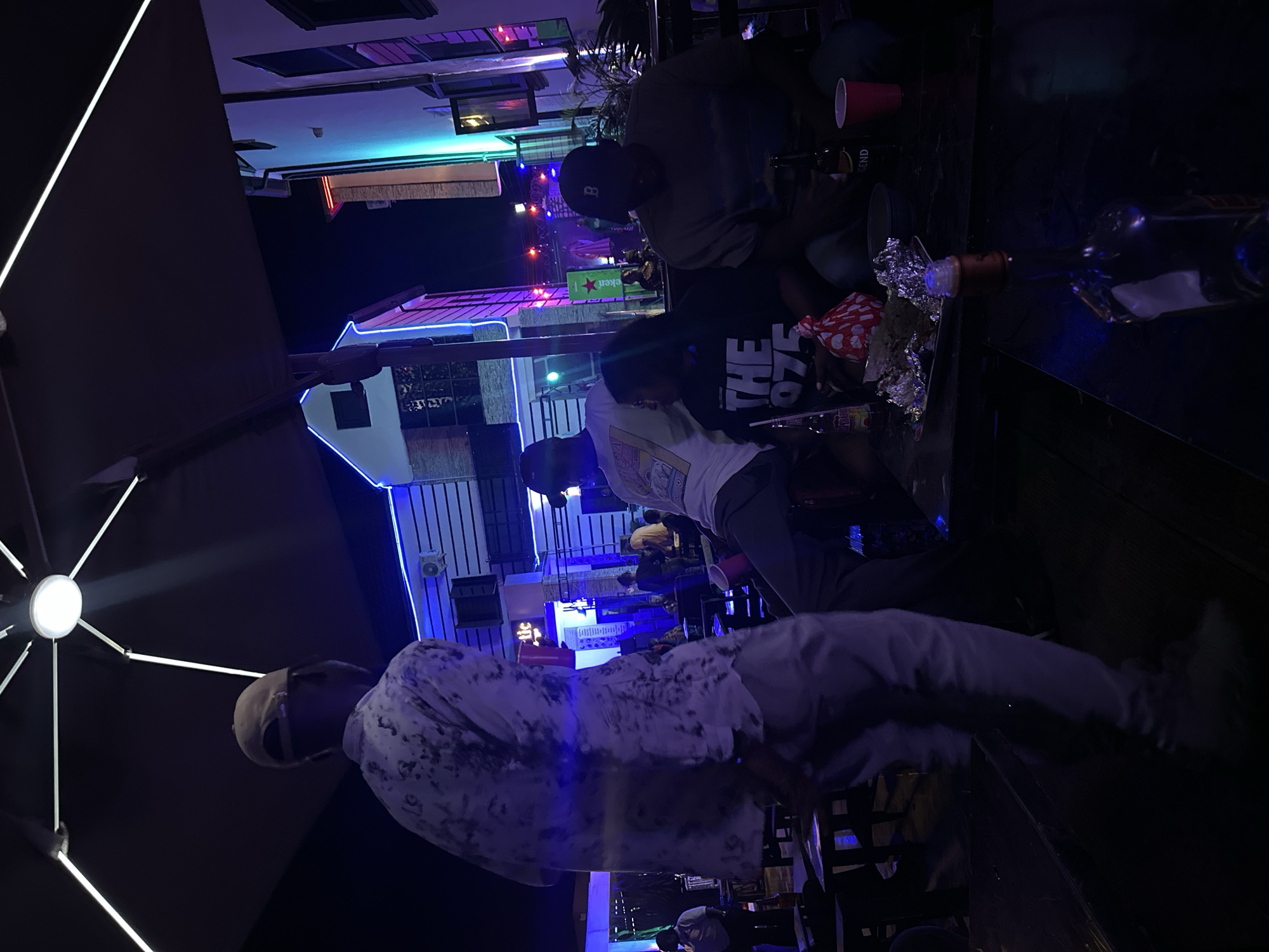 The image shows an outdoor gathering in Tumfure, Nigeria, at night. Three people are prominently visible around a table. The individual closest to the camera wears a light-colored patterned shirt and light pants. The person in the middle wears a white t-shirt with black lettering, while the farthest person wears a dark shirt and a baseball cap. There are several bottles and drinking vessels on the table. In the background, there is a building illuminated with colorful lights. A large umbrella or awning is partially visible in the foreground.