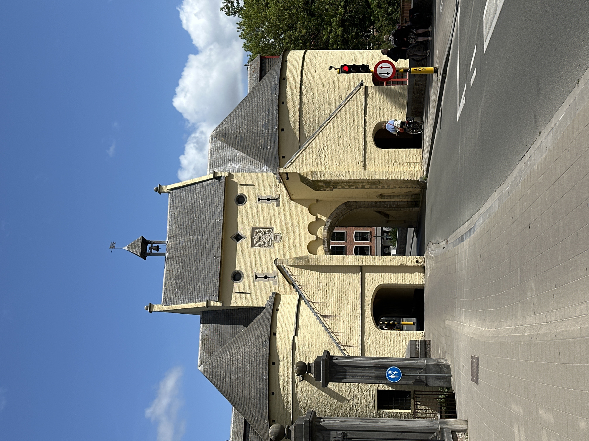 This image depicts a view of a historic yellow stone building with grey tiled roofs, characteristic of Bruges, Belgium. The building appears to be an old gate or part of a larger structure, with arched passageways and architectural details like small windows and decorative stonework. A small bell tower with a weather vane is visible on the left side of the building. The scene is bathed in bright sunlight, suggesting it is daytime, and the sky is a clear blue with a few fluffy white clouds.
A traffic light showing red is visible above the building, indicating traffic control. A sign with red arrows pointing in opposite directions, likely a traffic regulation sign, is also present. A person is seen riding a bicycle, emerging from one of the arched openings of the building and heading towards the street. In the background, there are trees and other people, suggesting a public area. A blue circular sign with a white silhouette of a person on a bicycle is mounted on a dark pillar in the foreground, indicating a bike path or pedestrian crossing.
The overall atmosphere seems peaceful and slightly touristy, typical of a sunny day in a historic European city. The architecture strongly points to a location like Bruges, known for its well-preserved medieval buildings and canals. No discernible text is visible in the image, other than what might be on the traffic signs, which are not clearly legible.