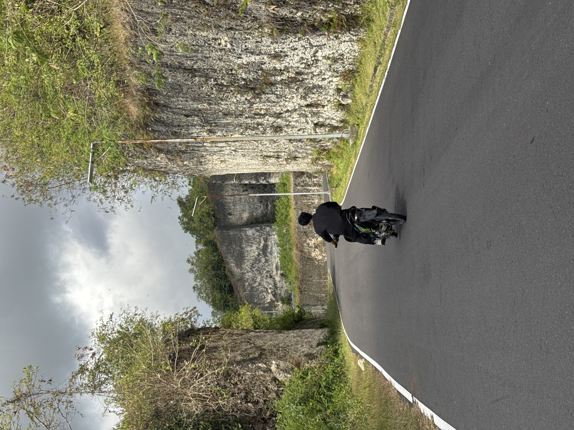 A lone individual rides a motorcycle down a paved road that cuts through a rocky landscape. The road is dark asphalt with a white line marking the edge on the left side. To the left of the road, a steep, rocky cliff face is covered with dense green foliage and dry branches. To the right, another, larger cliff face rises, its textured, light-colored rock interspersed with patches of green vegetation and some dry, wispy plants. A street lamp with a modern design is visible on the right, near the top of the cliff. Further ahead, the road curves, flanked by similar rocky formations and more greenery. The sky above is overcast with dark, dramatic clouds. The overall impression is one of a road winding through a natural, rugged environment under a moody sky. This scene is located in Kuta Selatan, Indonesia, a region known for its dramatic coastal cliffs and lush vegetation.