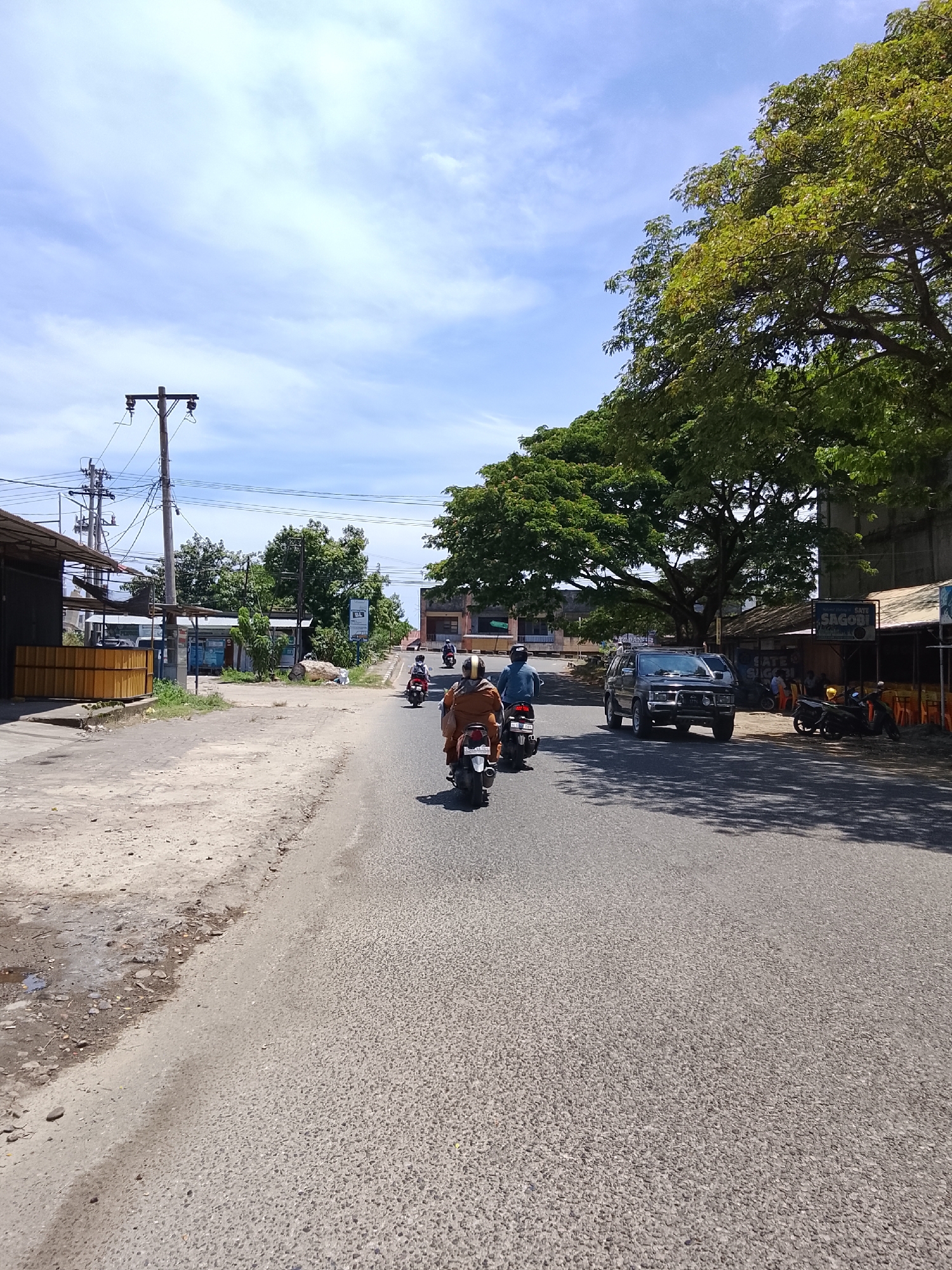 The image depicts a street scene in Lhokseumawe, Indonesia, under a bright, partly cloudy sky. In the foreground, a paved road occupies the majority of the frame, with a dirt shoulder on the left. Several motorcycles are traveling on the road, with riders visible on each. One motorcycle in the immediate foreground has a rider wearing an orange top. Further down the road, a dark-colored SUV is in motion.
Along the sides of the road, there are various buildings and structures. On the left, a dark-colored building with a yellow structure in front of it is visible, along with utility poles and wires. On the right, there are open-fronted shops or stalls, some with seating and awnings, and signs including one that reads "Sate Sagobi". A large, leafy tree with a wide canopy dominates the right side of the scene, casting shadows on the road. The environment appears to be a commercial or mixed-use area within the city.