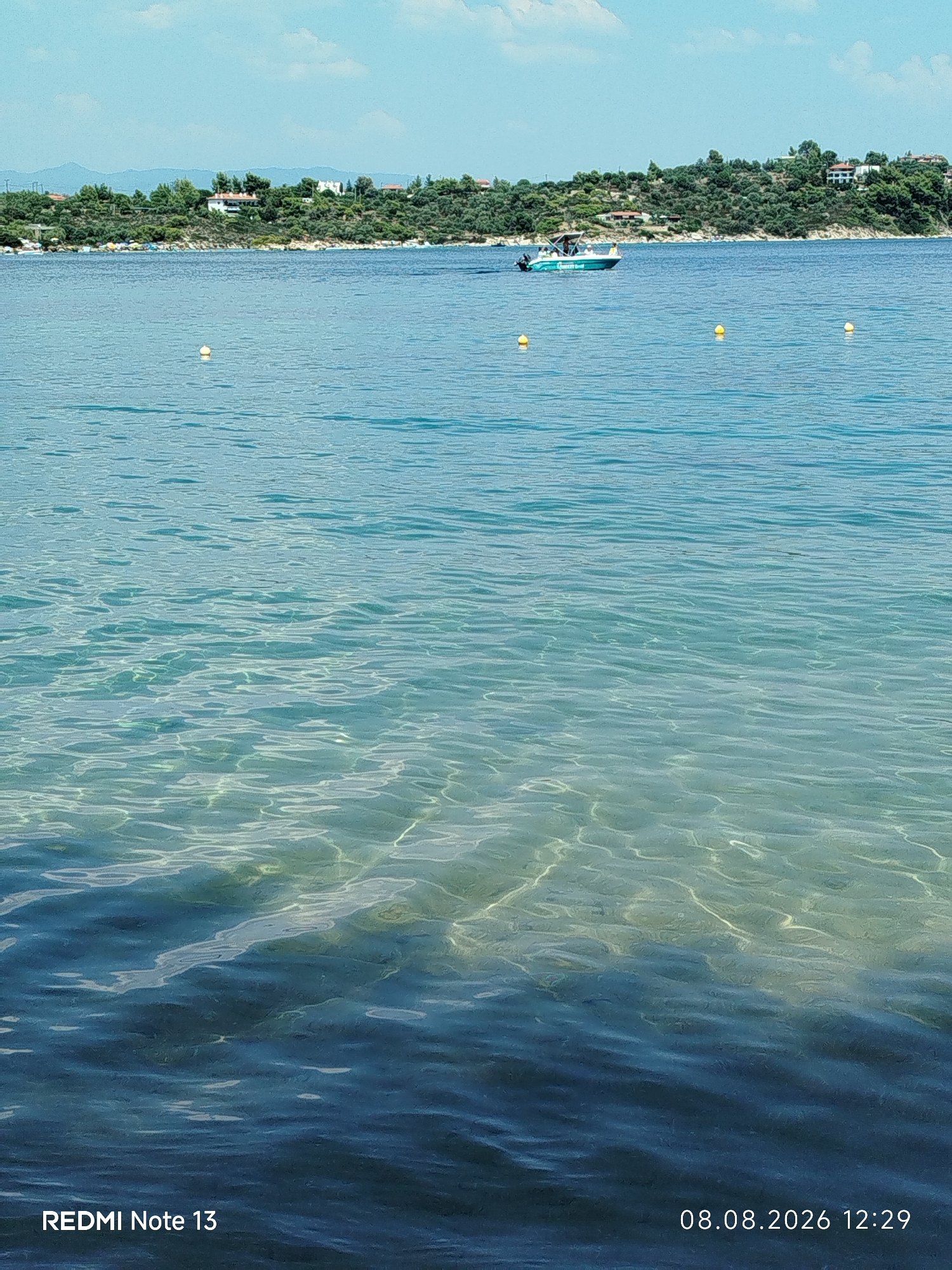 This is a landscape photograph of a coastal scene in the Sithonia Municipal Unit, Greece. The foreground is dominated by the clear, blue water of the sea, with sunlight reflecting off the rippling surface. The seabed is visible in some areas, appearing sandy and light-colored. In the mid-ground, a few yellow buoys are scattered across the water. A small boat with a canopy and an outboard motor is moving away from the shore. The background features a densely vegetated coastline with scattered houses and trees. The sky is a pale blue with some wispy clouds. The bottom left corner of the image displays "REDMI Note 13" and the bottom right corner shows "08.08.2026 12:29".
