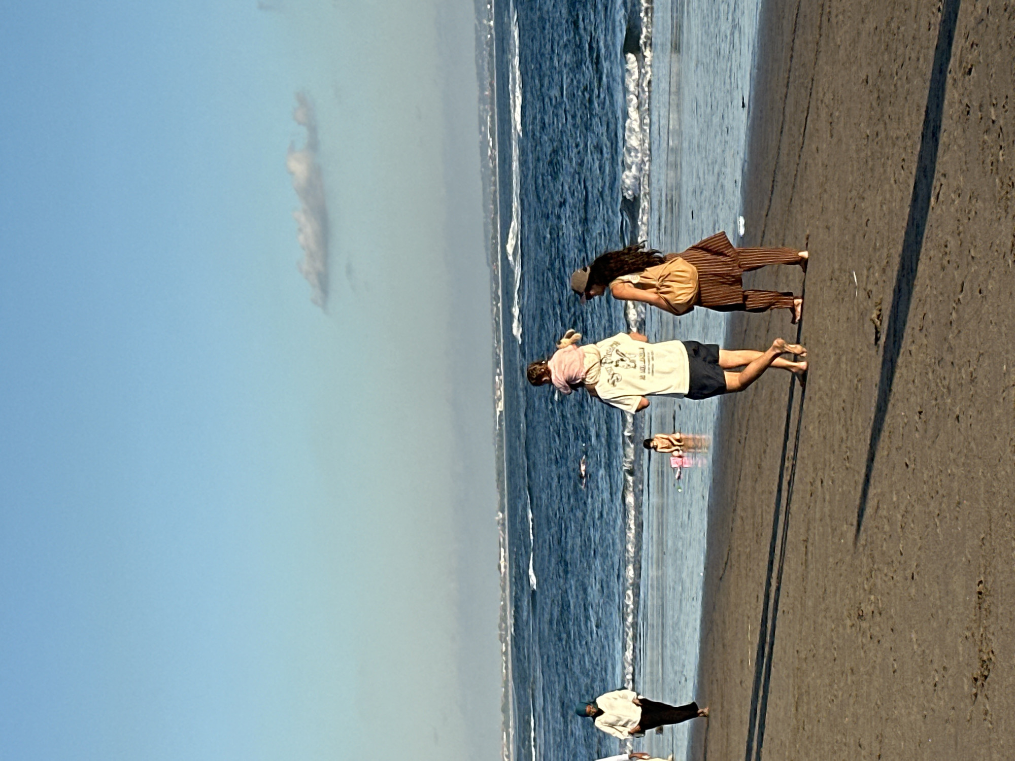 On a sandy beach in North Kuta, Indonesia, a group of people walk near the ocean. The sky is a pale blue with a single wispy cloud. The ocean is a deep blue with white-capped waves. In the foreground, a man carries a toddler on his shoulders, and a woman walks beside him. To the left, another woman walks along the beach. In the background, a child sits in the shallow water. The overall scene depicts a relaxed day at the beach.