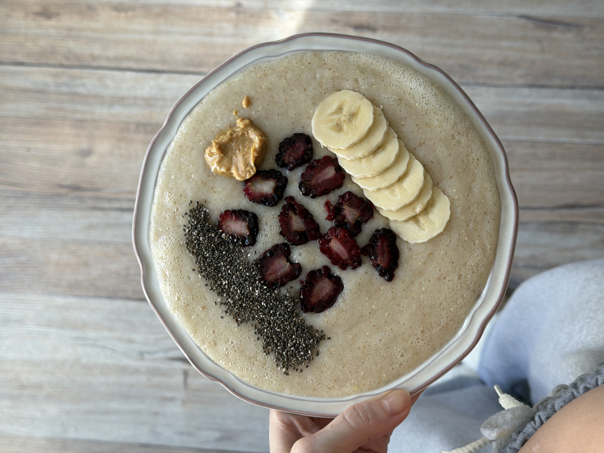A top-down shot reveals a hand holding a white decorative plate of smoothie. The smoothie is light brown and topped with chia seeds, peanut butter, sliced bananas, and cut dark-red fruit. The background is a light-brown wooden-plank-patterned surface. The location is not discernible from the image.