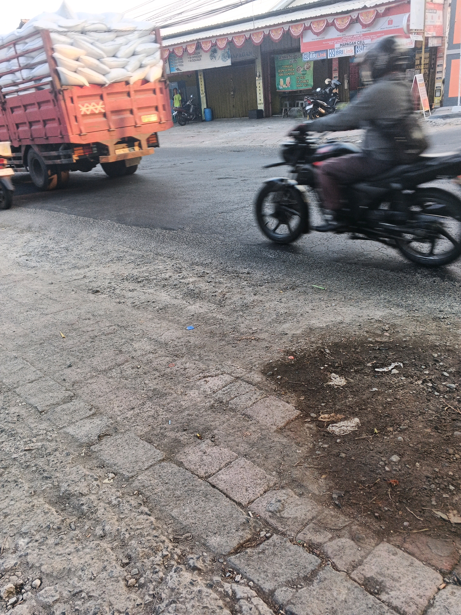 The image shows a street scene in Ungaran, Indonesia. A large red truck carrying bags of what appears to be rice is driving on the road, partially obscuring a man on a motorcycle who is also in motion. The truck is on the left side of the frame, and the motorcycle is in the center-right. The road is paved with asphalt and has some gravel and dirt on the shoulder. There are buildings with awnings and signage visible in the background. One sign reads "Mapagan Cell". The lighting suggests it is daytime, with some shadows present.